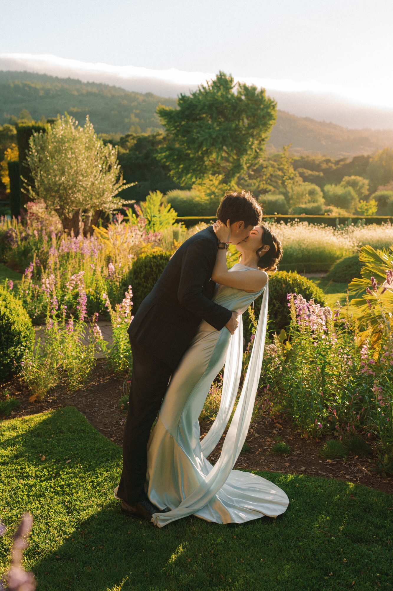 A couple in wedding attire sharing a kiss outdoors with trees and mountains in the background.