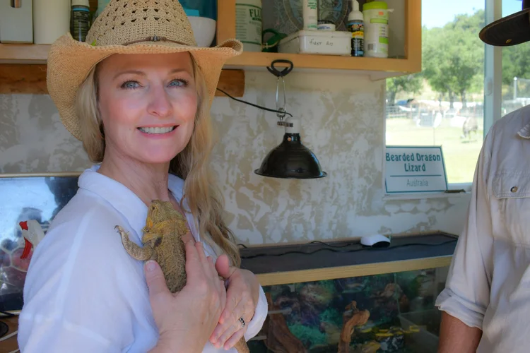 Woman with blonde hair wearing a straw hat and white shirt, smiling and holding a small animal, in a room with shelves and a window.