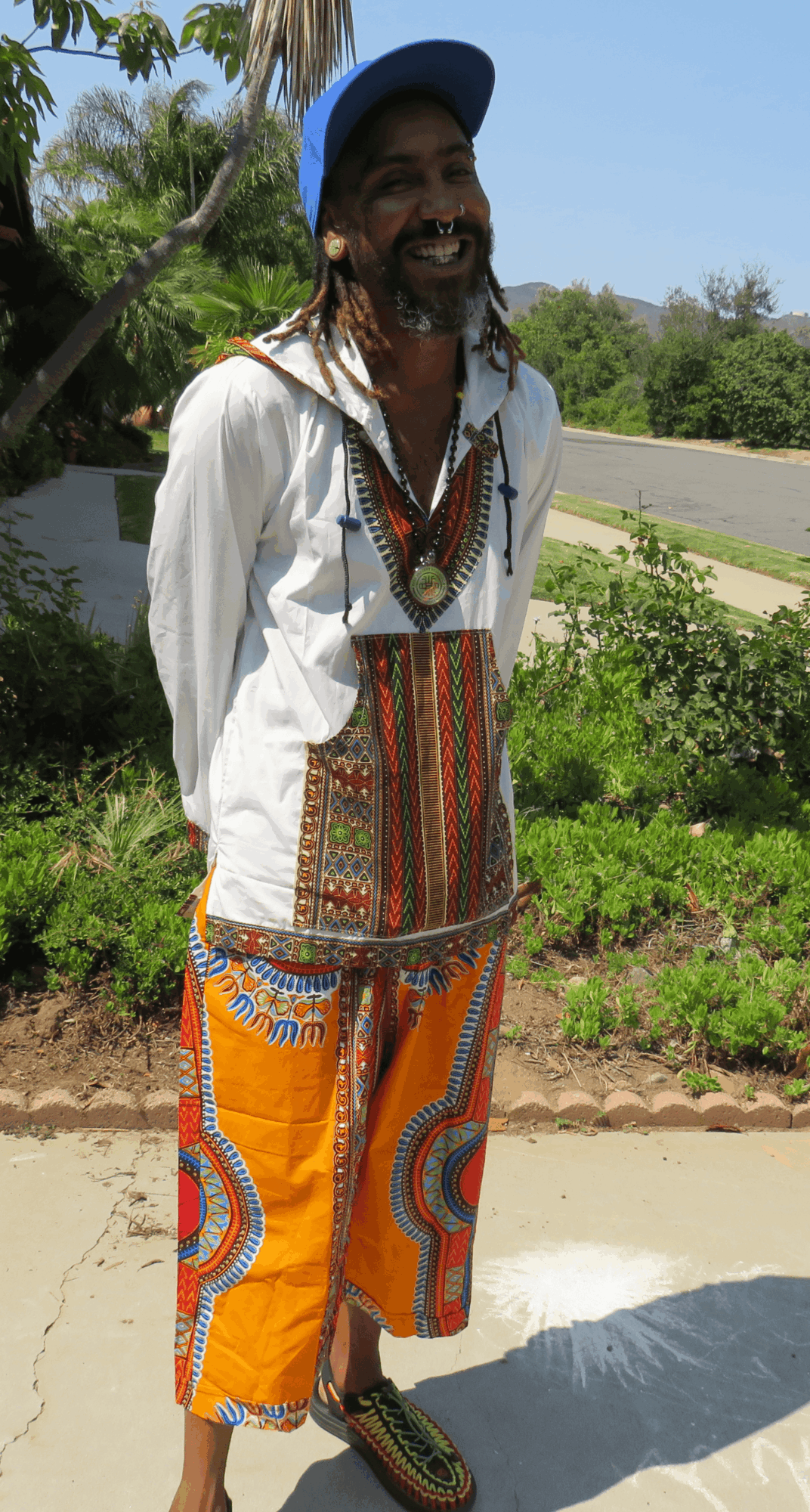 Smiling man with dreadlocks wearing colorful traditional clothing and a blue hat outdoors, sunny day, greenery in background.
