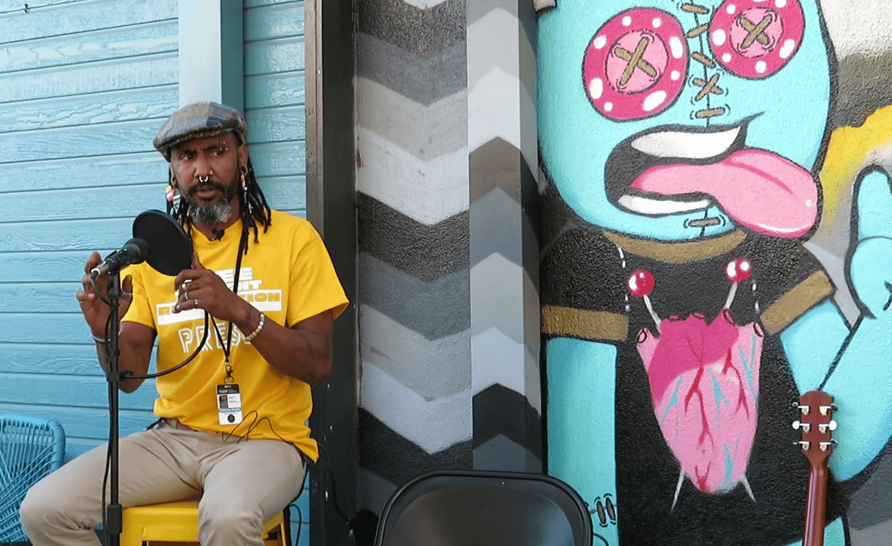 Man with dreadlocks wearing a yellow shirt and hat, sitting and holding a microphone, next to colorful street art of a cartoon character with glasses and tongue out.