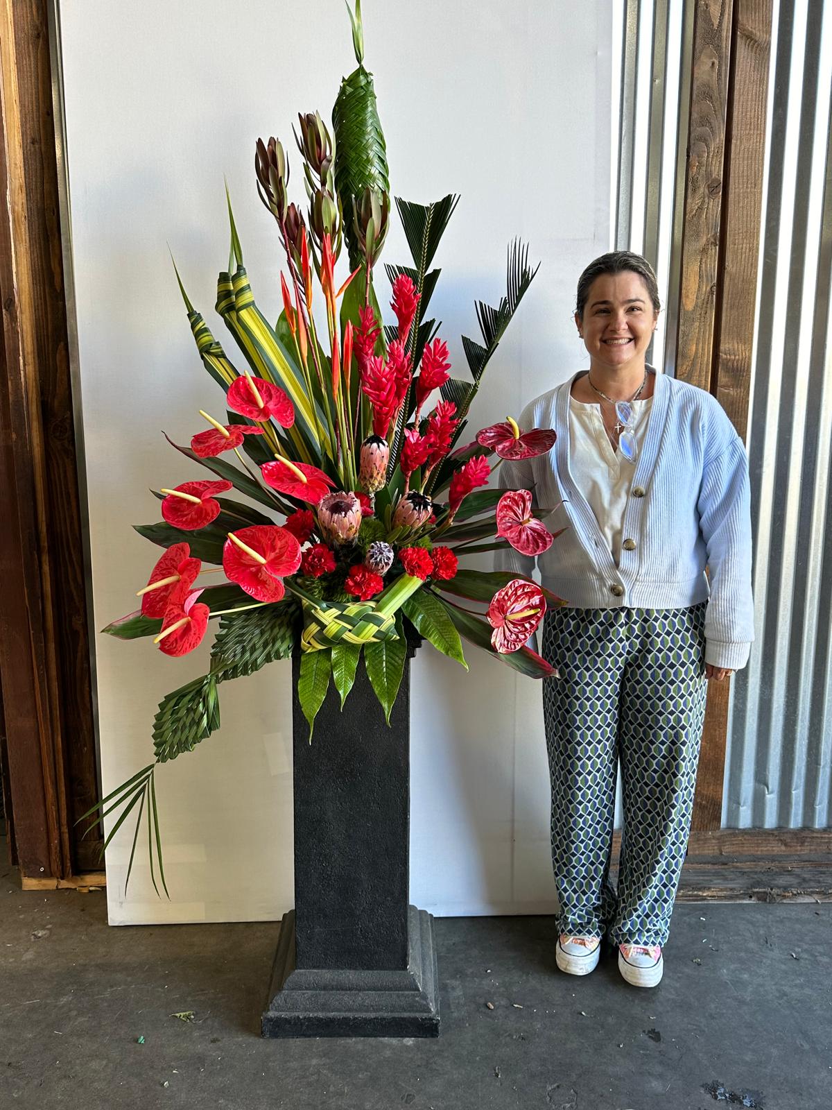 Woman standing next to a large floral arrangement in a black vase, smiling, indoors with a wooden floor and window.