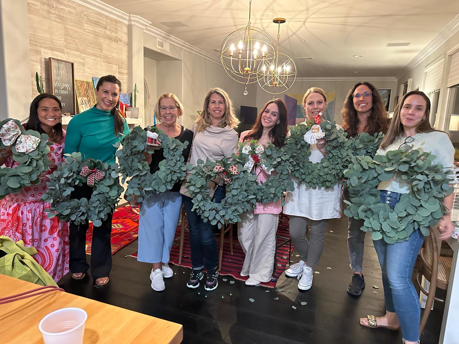 Group of eight women and one man holding large green wreaths with pink flowers, standing indoors in a decorated room.