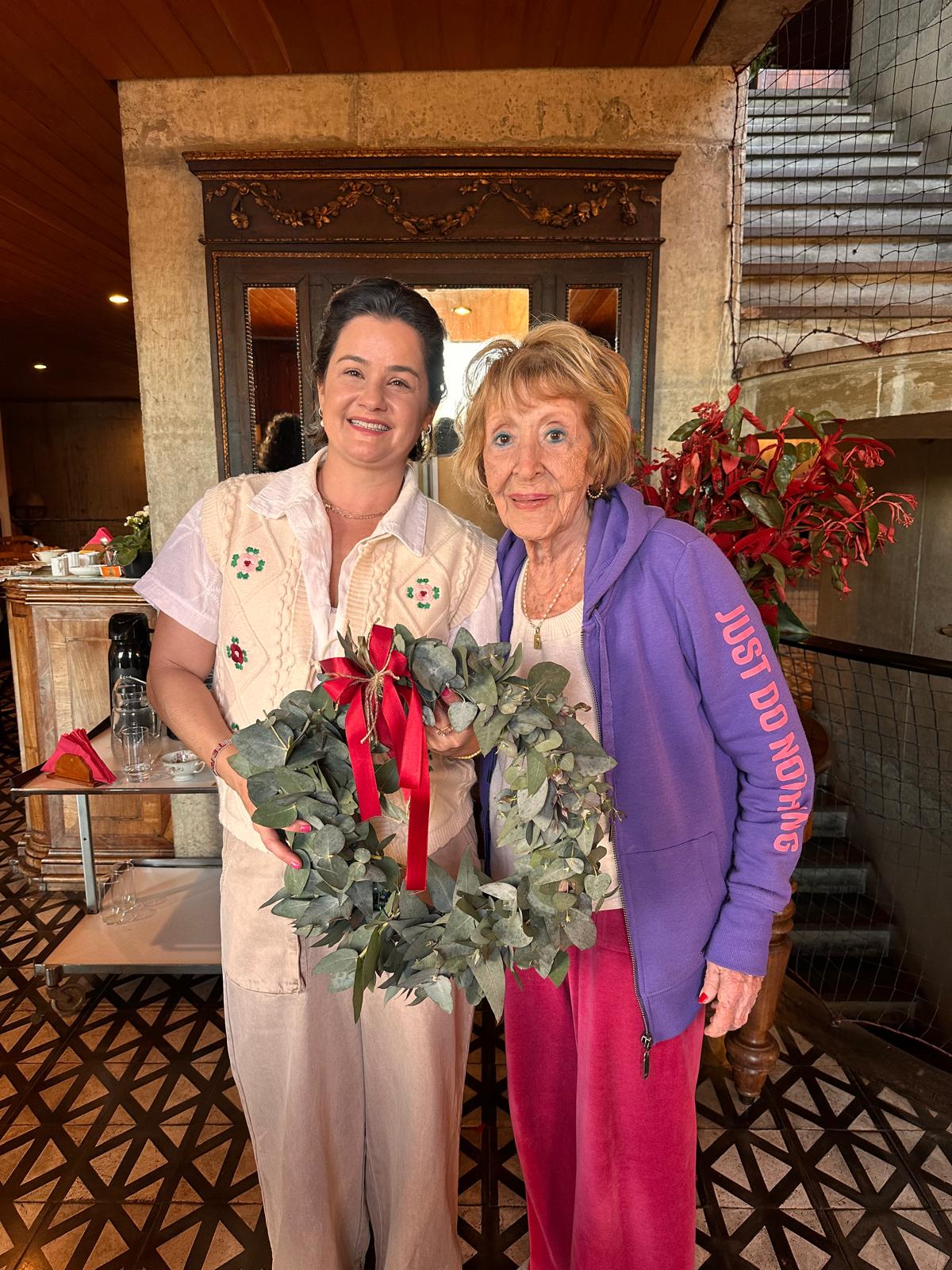 Two women standing indoors, holding a Christmas wreath with red ribbon, smiling, with a staircase and plants in the background.