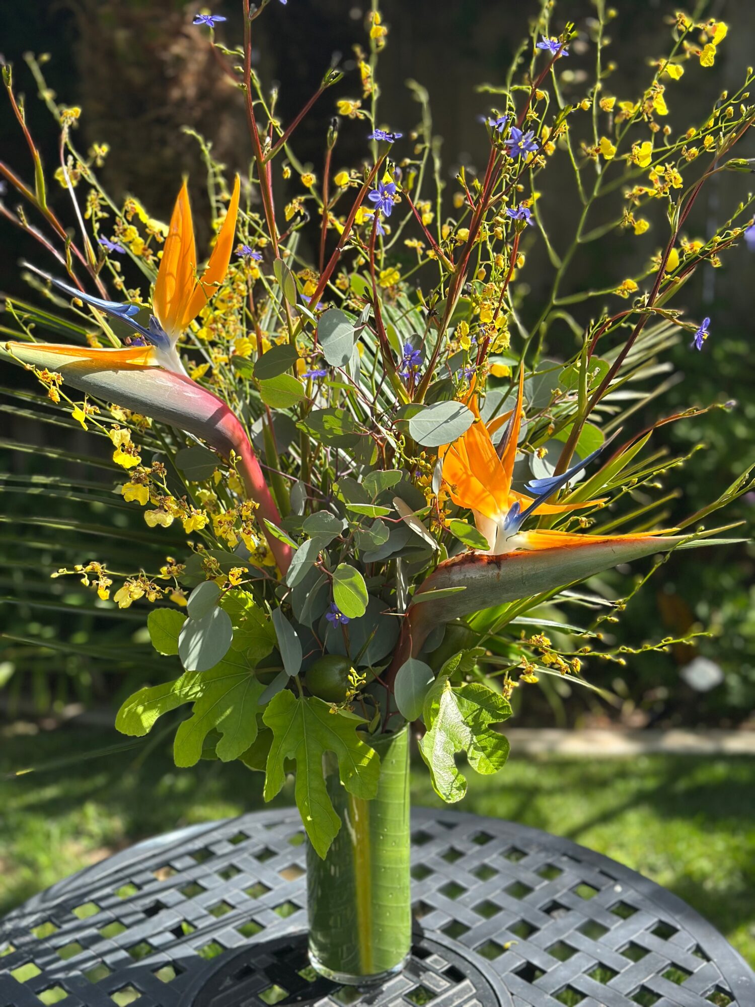 Vase with colorful flowers and green leaves on a table outdoors, sunlight in background.