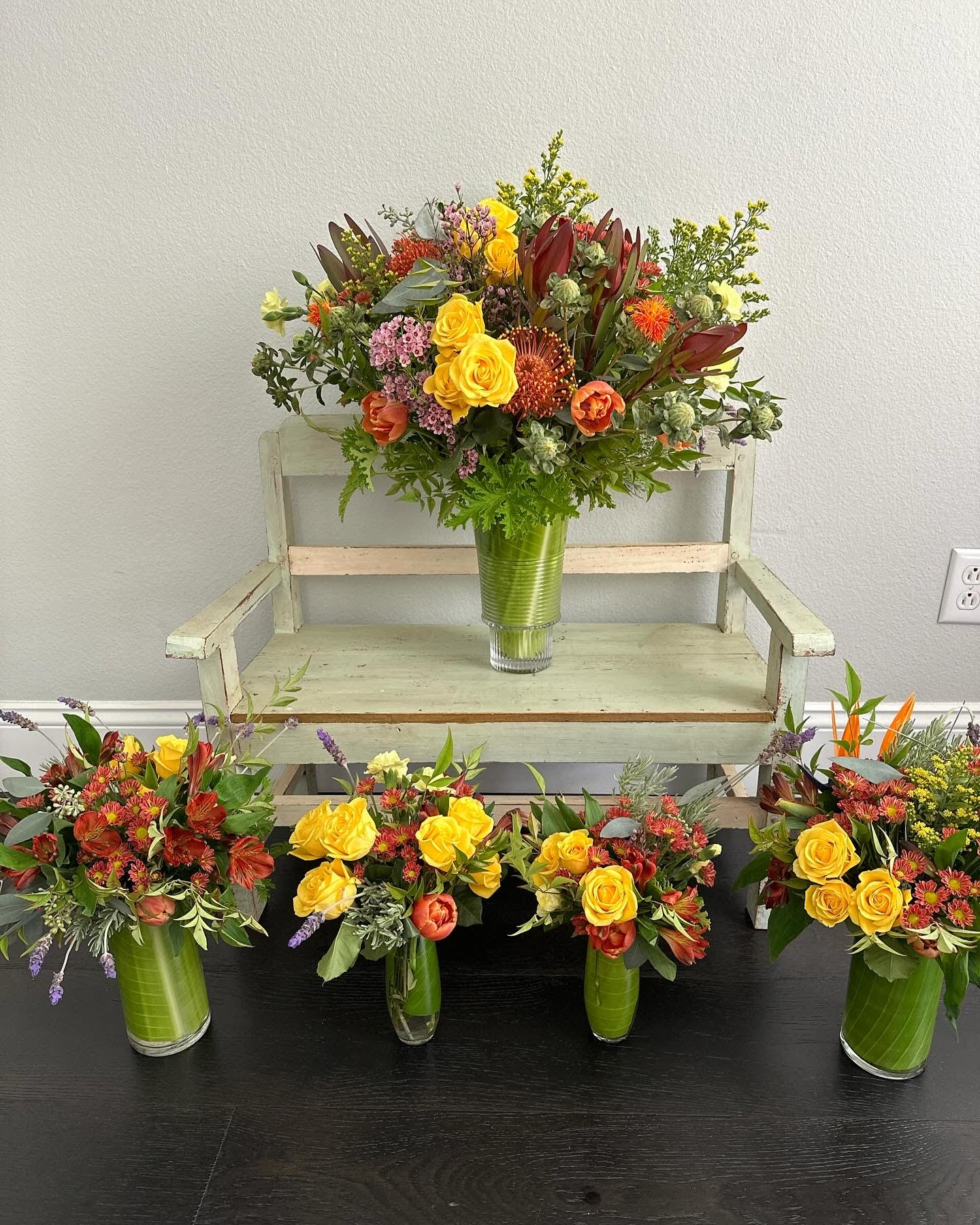 A large floral arrangement in a green vase on a white bench, with four smaller arrangements on the floor, all with colorful flowers.