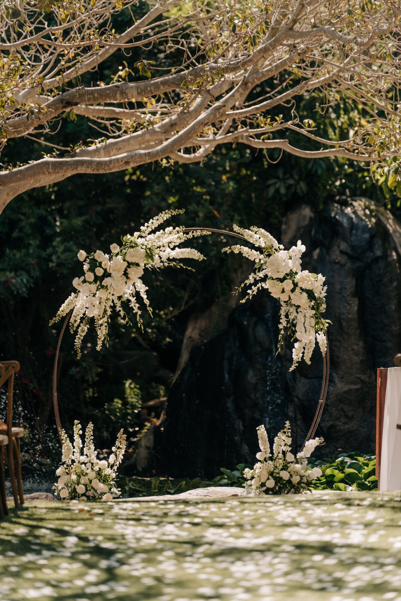Decorative floral arch with white flowers and greenery outdoors, with trees and rocks in background.