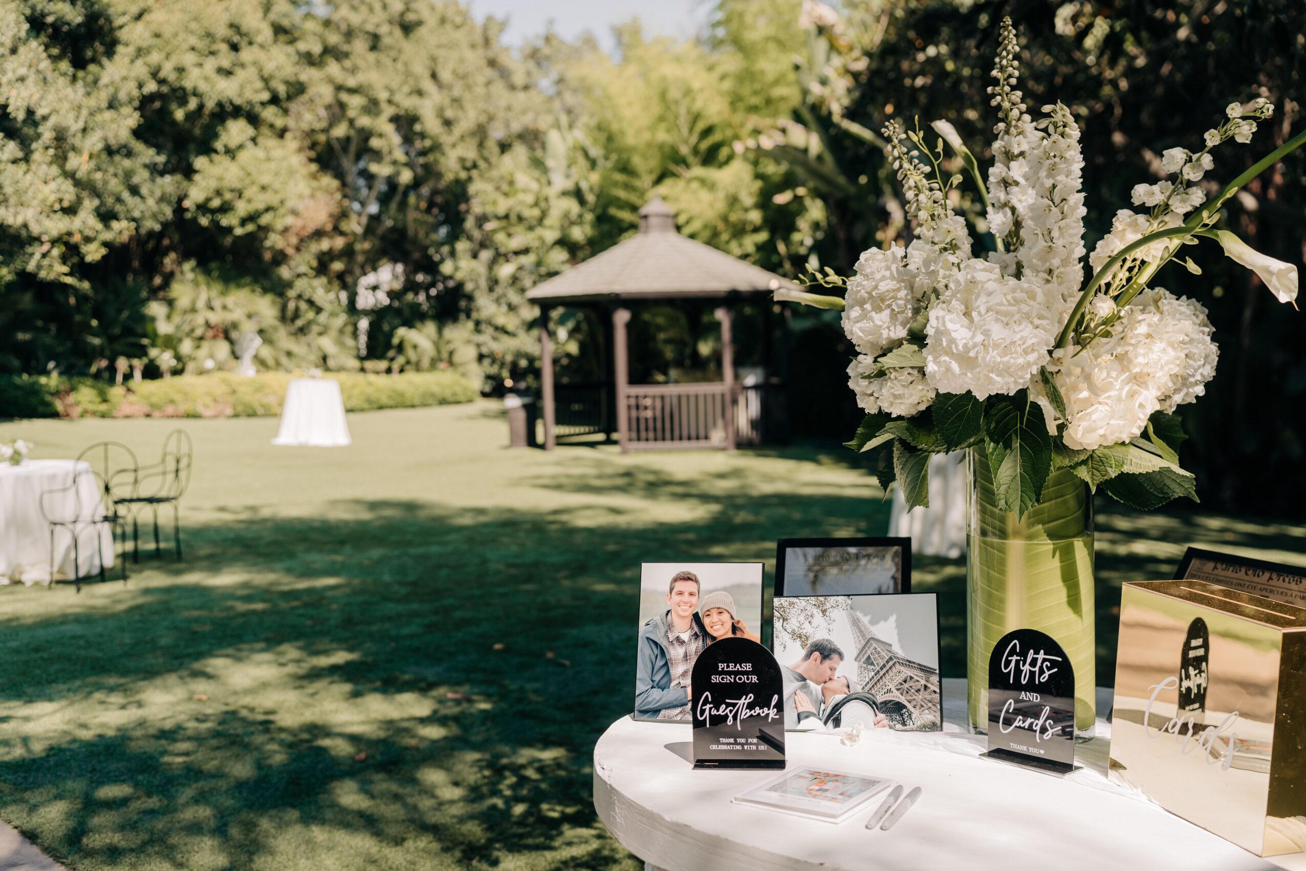 Table with photo frames and decorations outdoors, with a gazebo and trees in the background, and a large flower arrangement nearby.