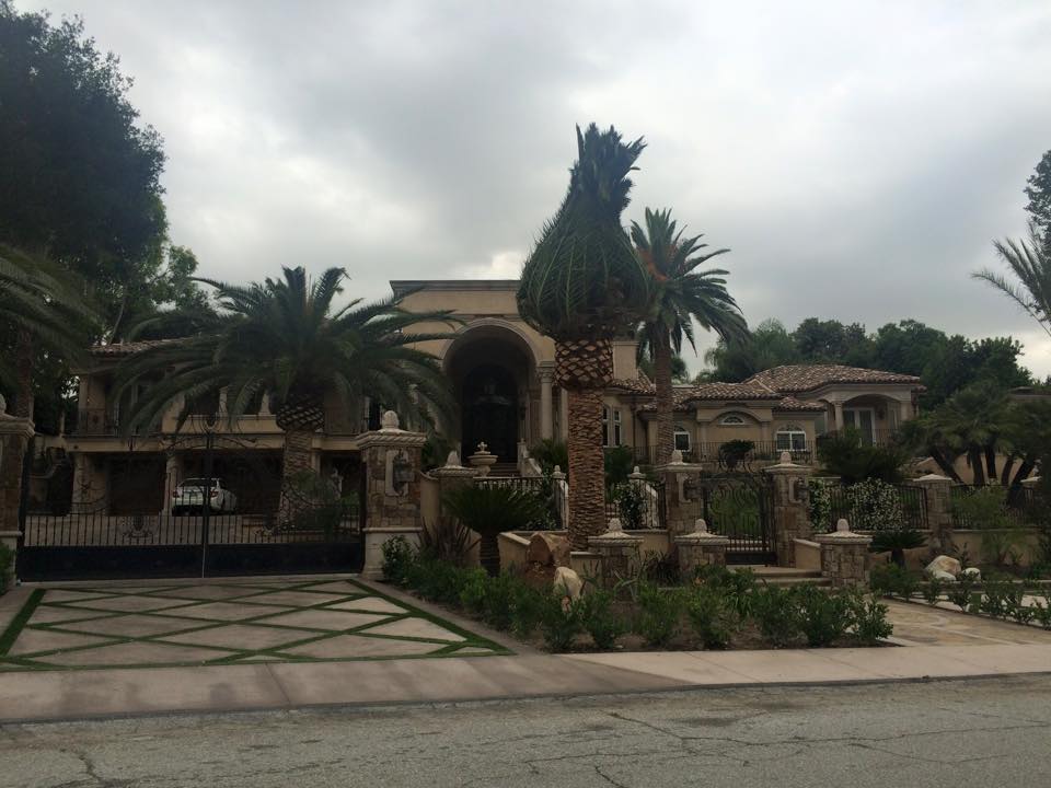 Large house with arched entrance, surrounded by palm trees and landscaped garden, under cloudy sky.