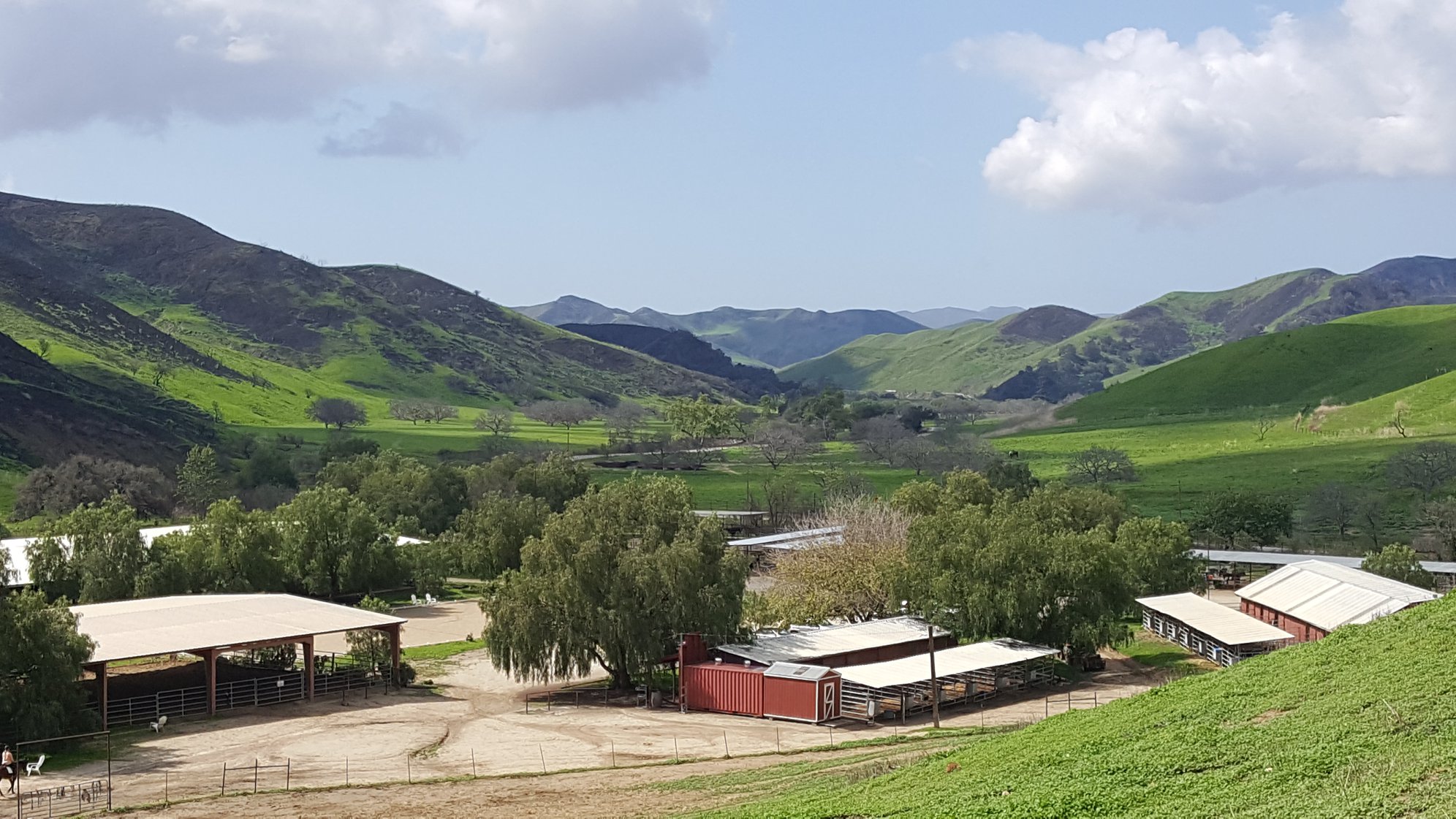 Rural landscape with green hills, trees, and farm buildings under a partly cloudy sky.