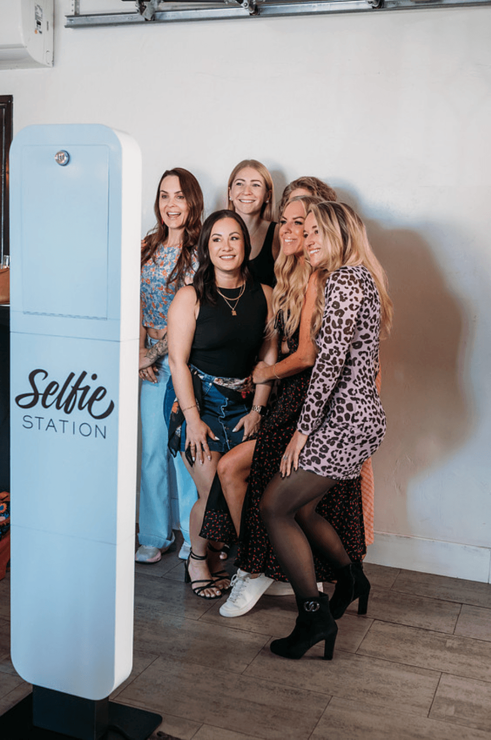 Group of five women standing together indoors near a Selfie Station, smiling and posing for a photo.