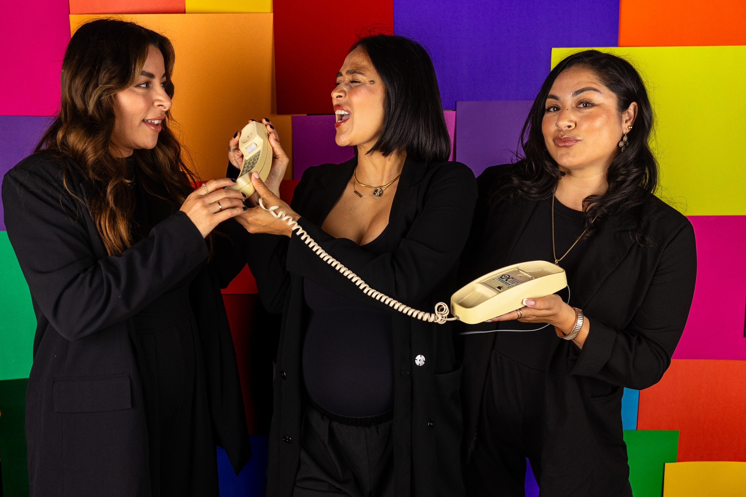 Three women in black clothing talking and smiling against a colorful, geometric background, one holding a vintage telephone receiver.