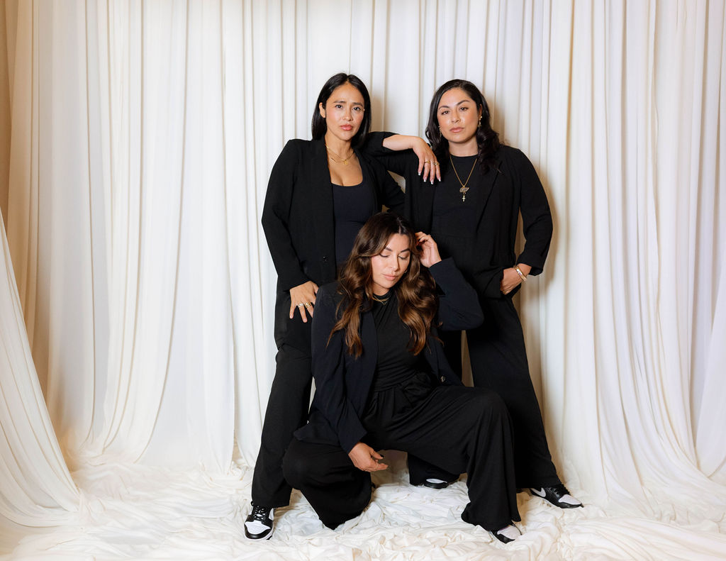 Three women in black outfits pose in front of white curtains, one sitting and two standing, with relaxed expressions.