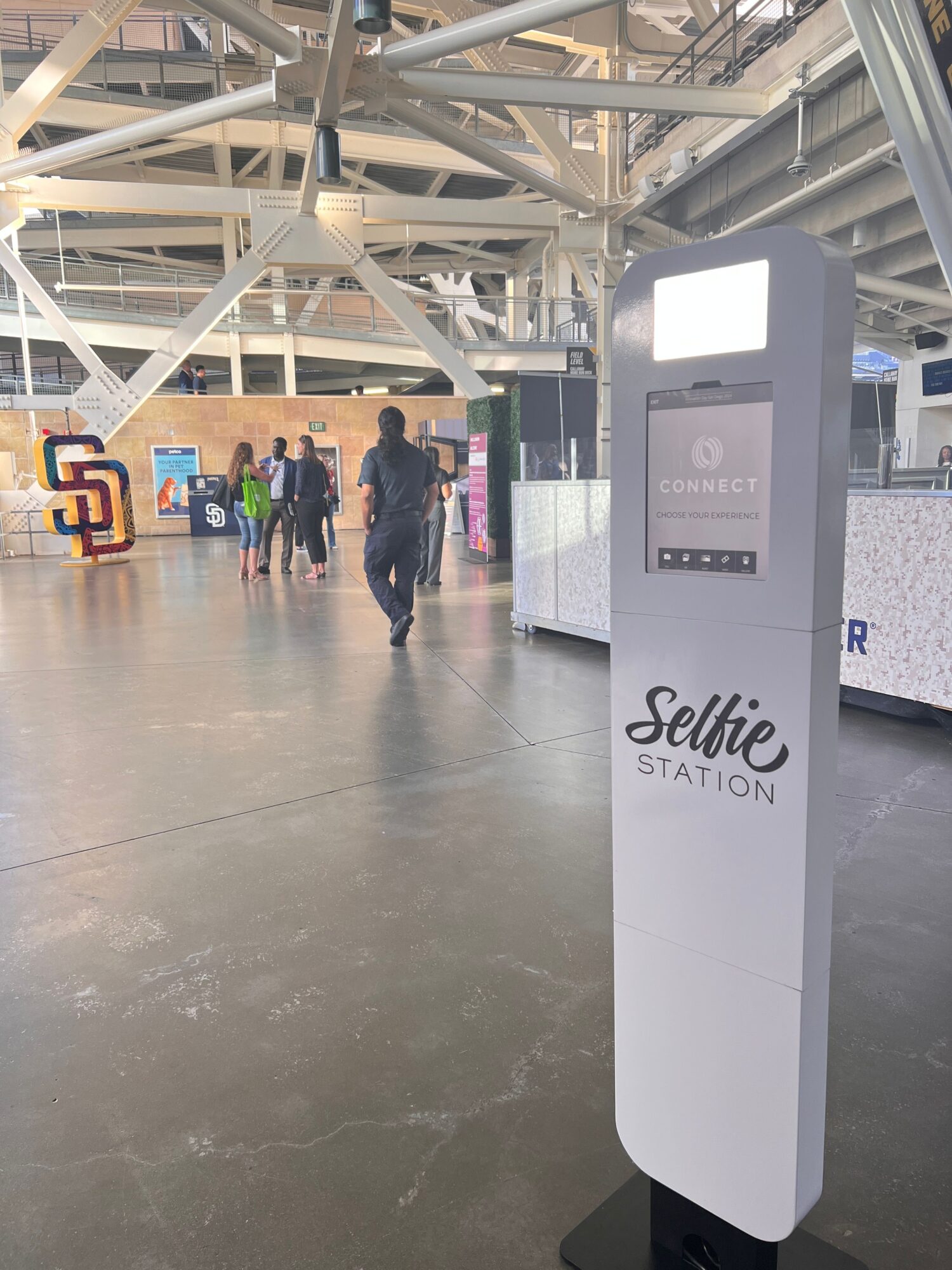 People walking in an airport terminal near a Selfie Station kiosk, with structural beams overhead.
