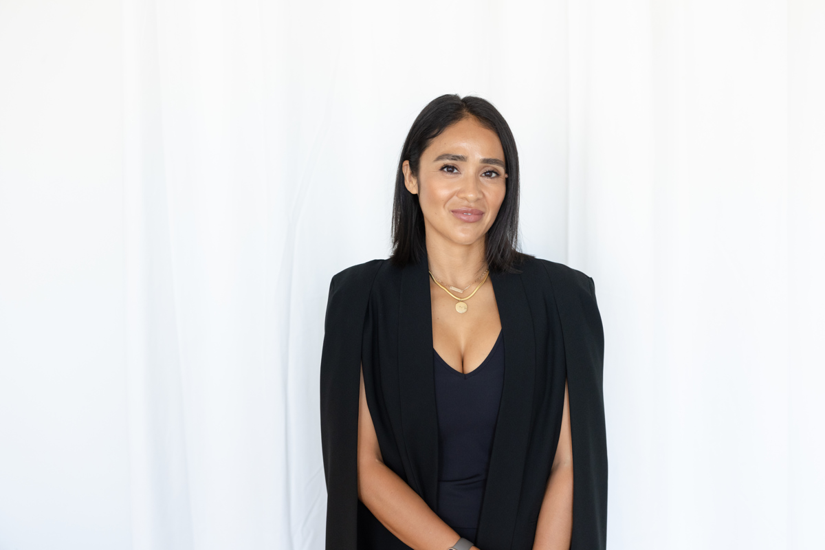 Woman with long dark hair wearing a black blazer and navy top, standing against a white background.