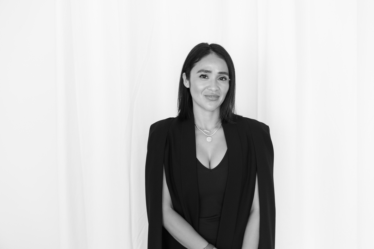 Woman with long dark hair wearing a black blazer and top, standing in front of a white curtain background.