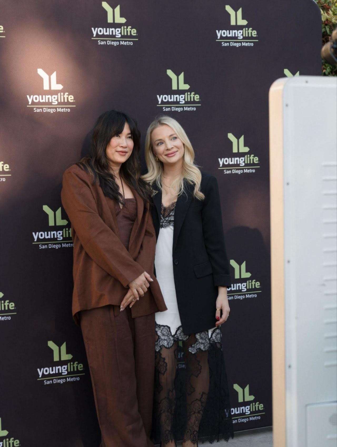 Two women standing together in front of a Young Life backdrop, smiling, one in brown suit, the other in black and lace dress.