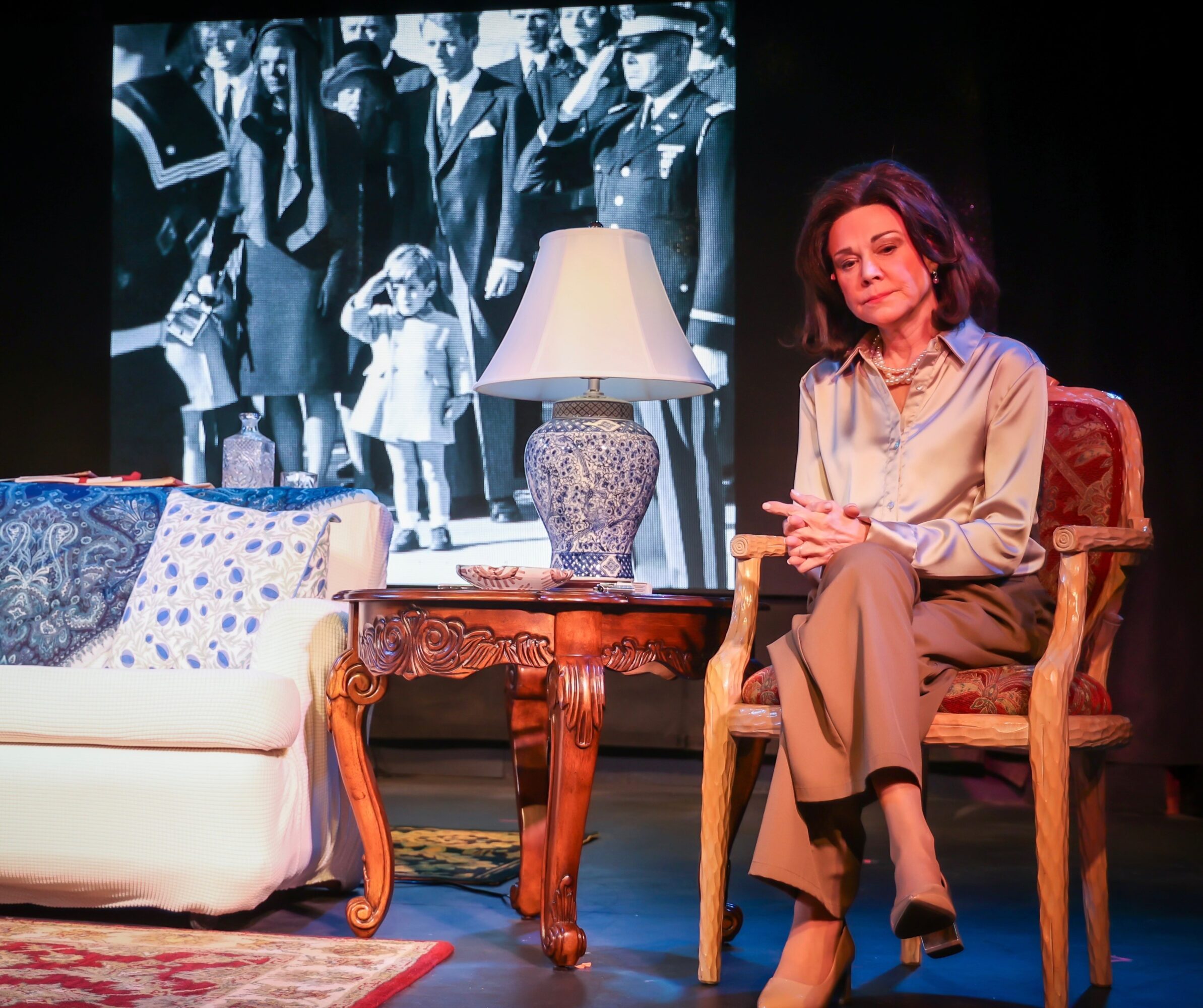 Woman sitting on a chair with a lamp and sofa, black-and-white photo in background, stage setting, 20 words