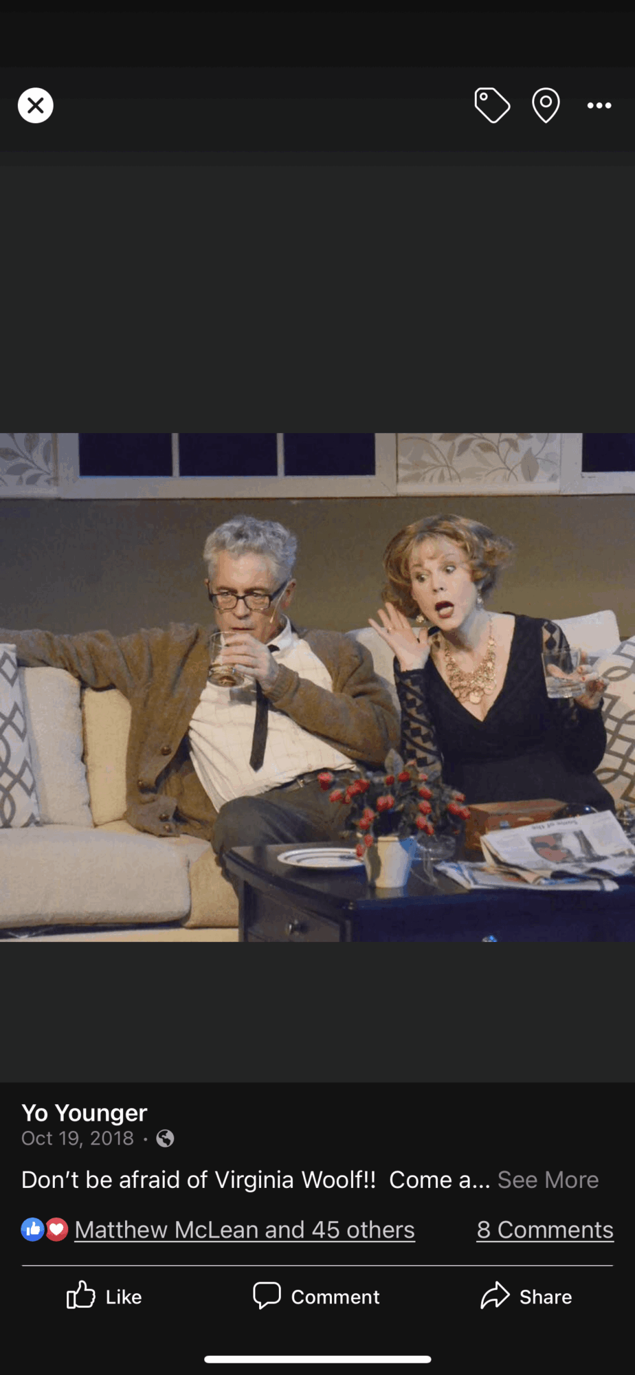 Two people sitting on a couch, holding cups, with a table in front, in a living room setting.