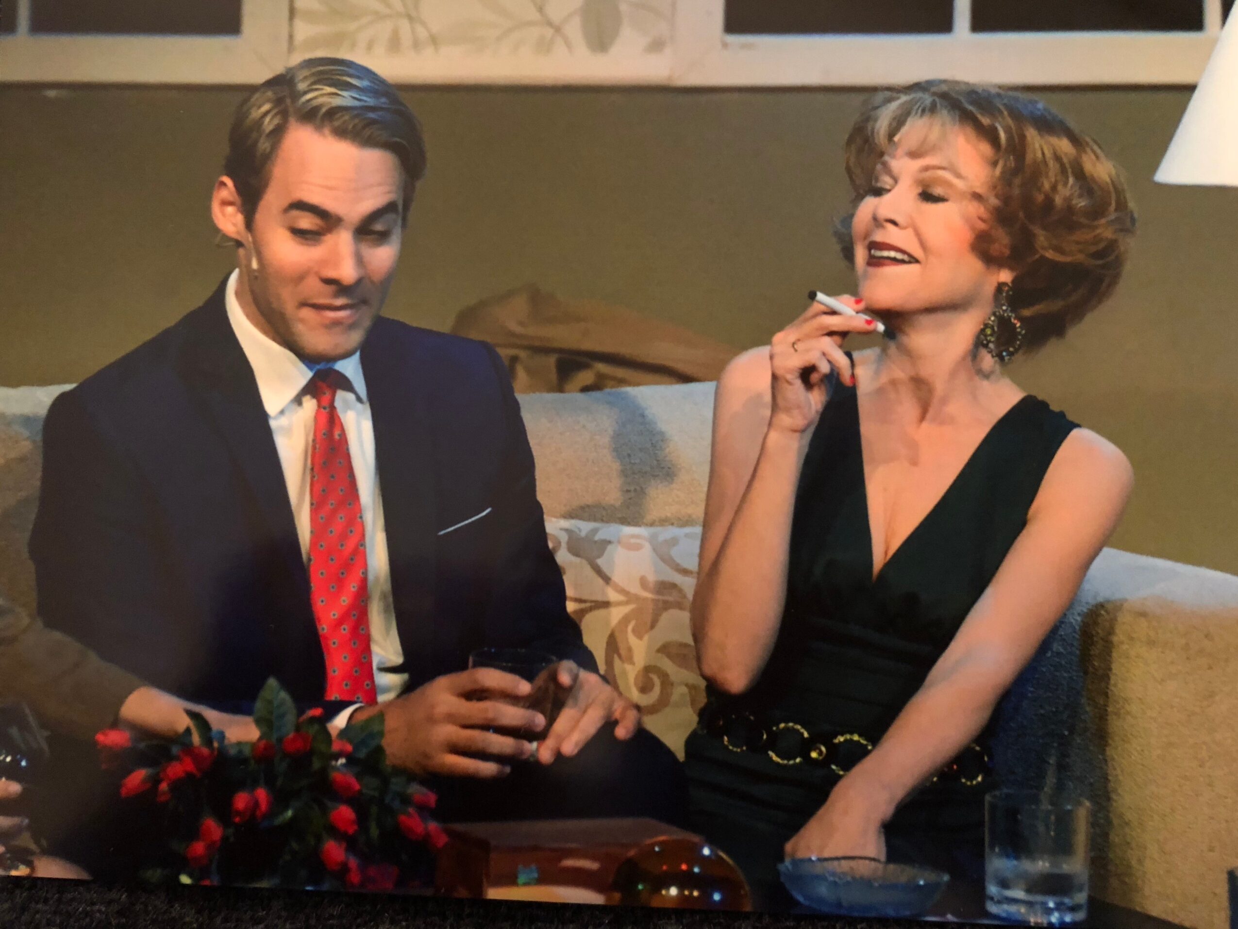 A man in a suit and a woman in a black dress sit on a sofa, with a glass of water nearby.