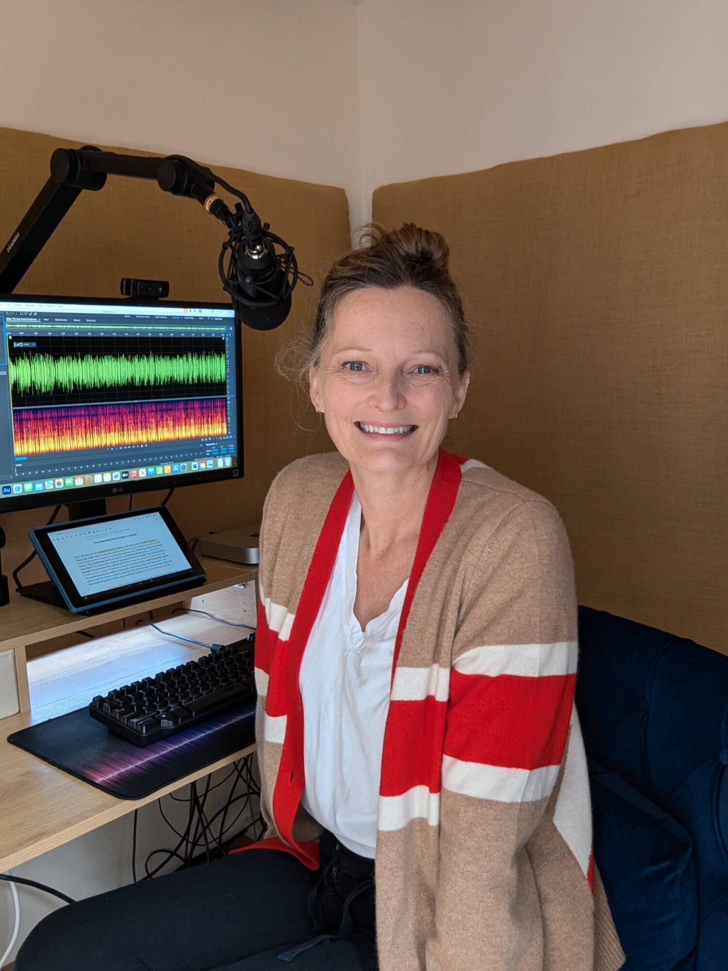 Smiling woman sitting at a desk with audio editing equipment and a computer monitor, in a recording studio.