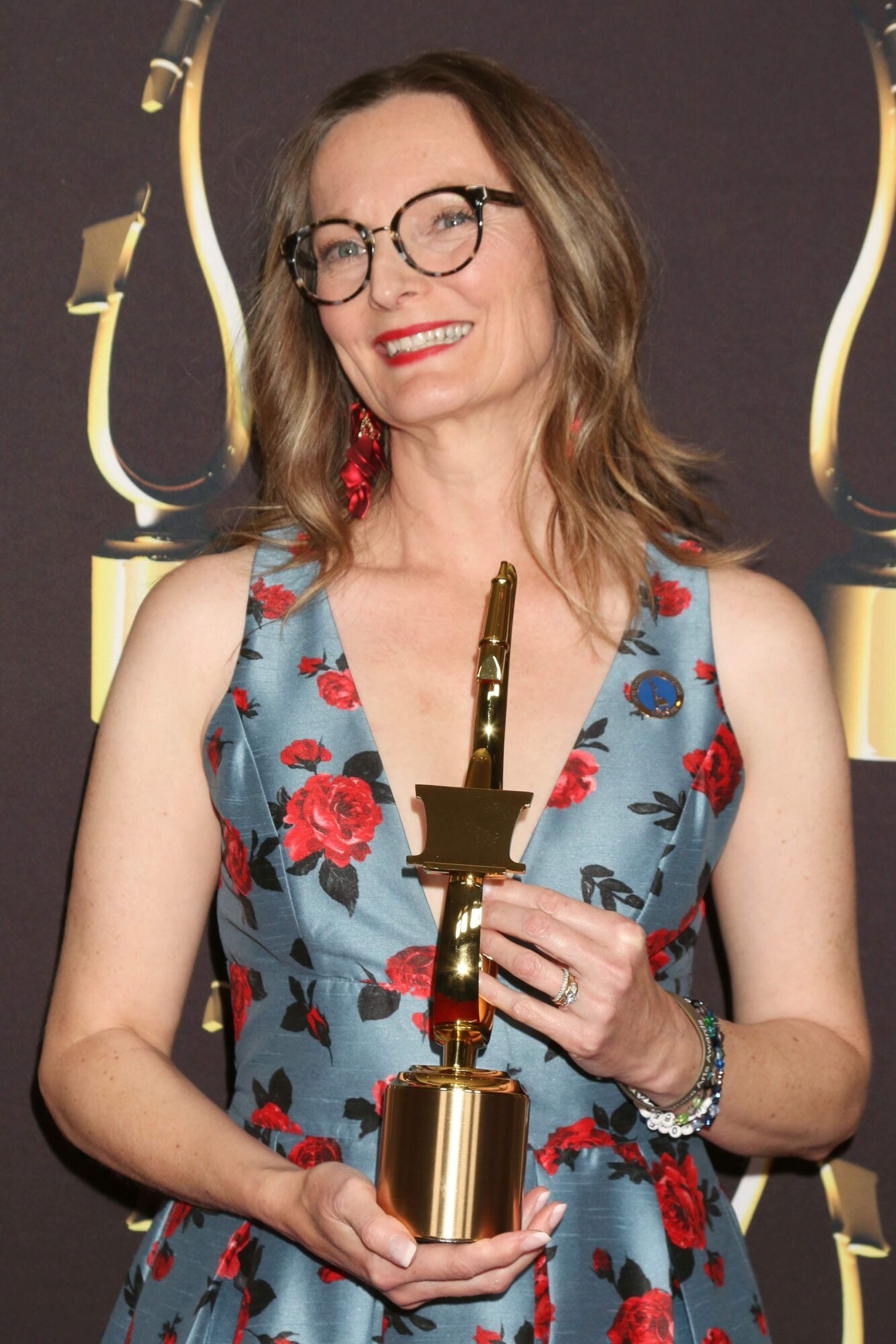 Woman with glasses in a floral dress holding an award trophy, smiling, dark background with gold accents