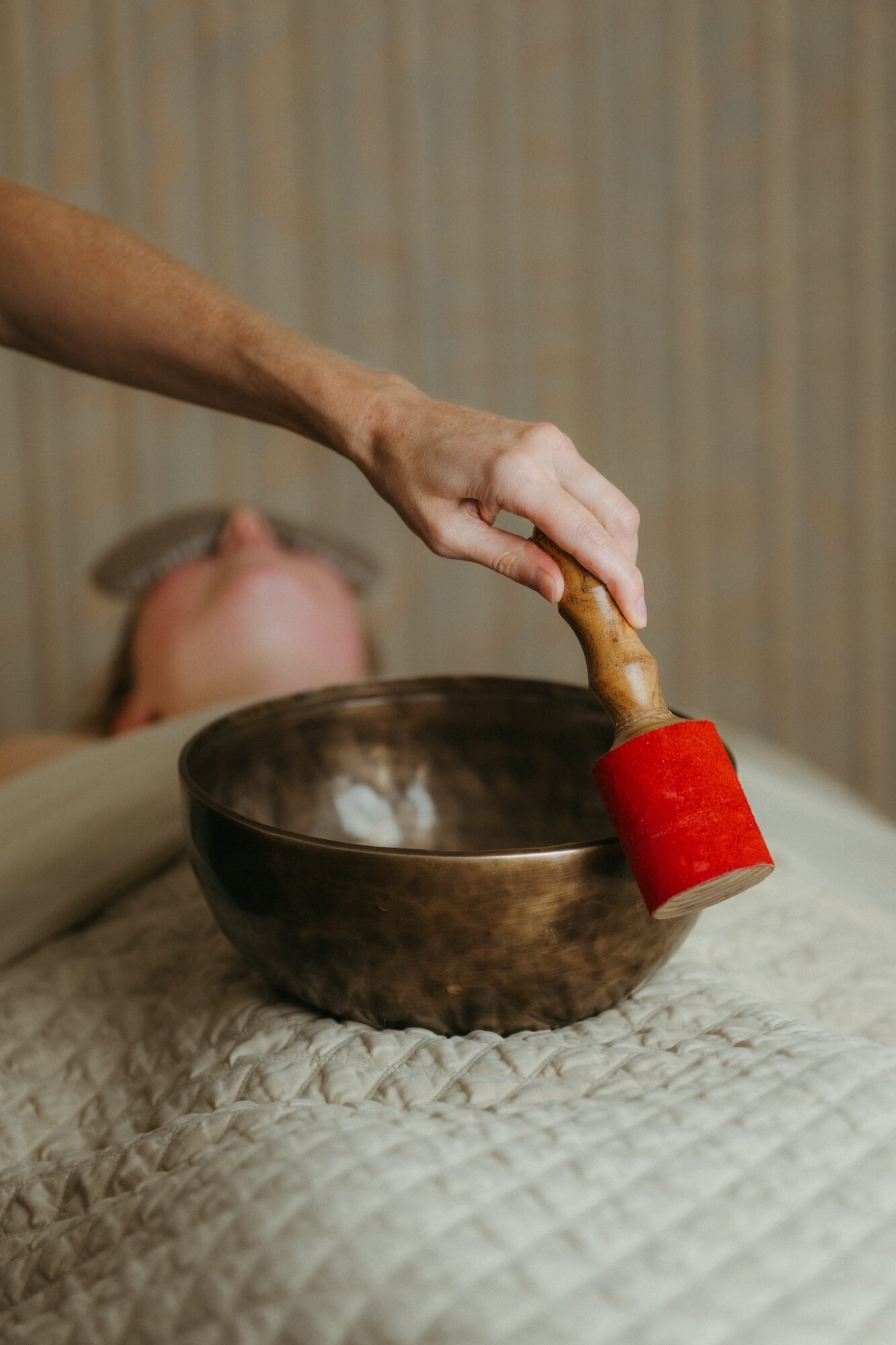 Person's hand holding a wooden massage hammer above a bowl, with a person lying down in the background, blurred.