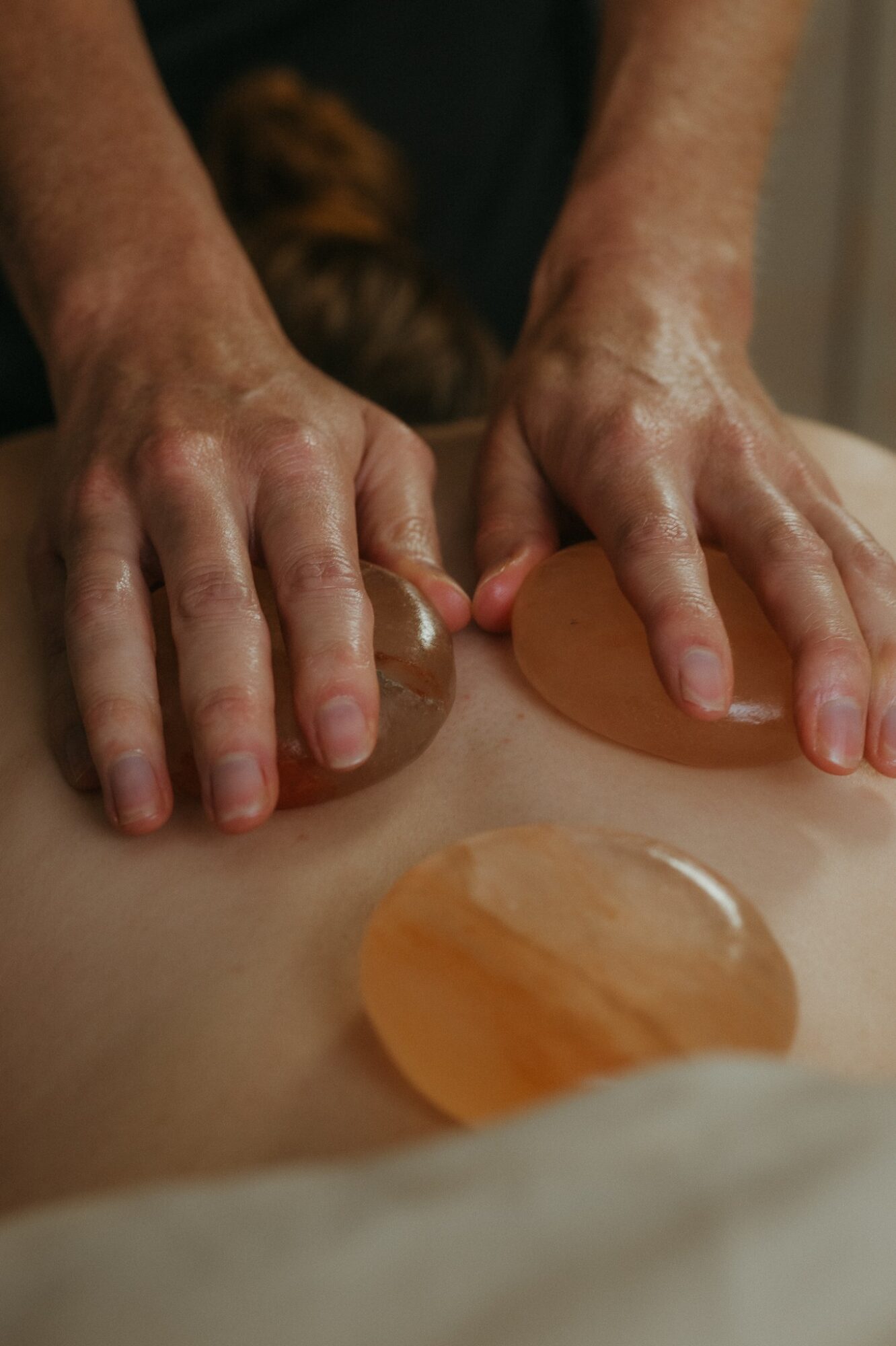 Hands of a massage therapist applying pressure on a person's back with hot stones, with additional stones nearby.
