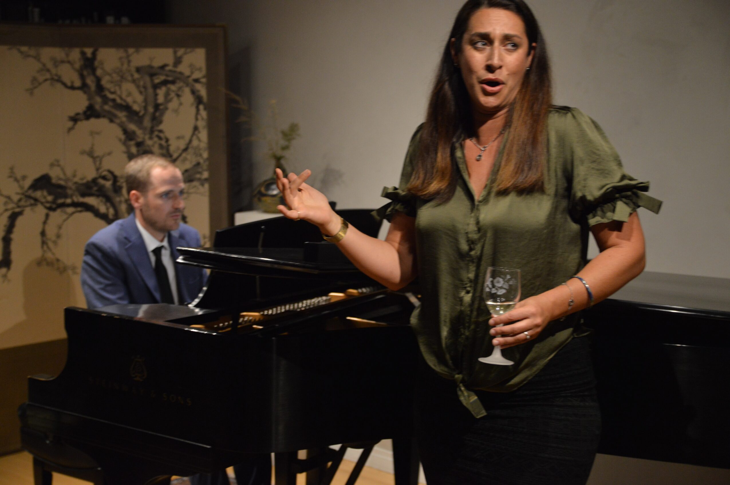 Woman speaking with a glass in her hand, man playing piano in background, indoor setting, neutral wall, framed artwork