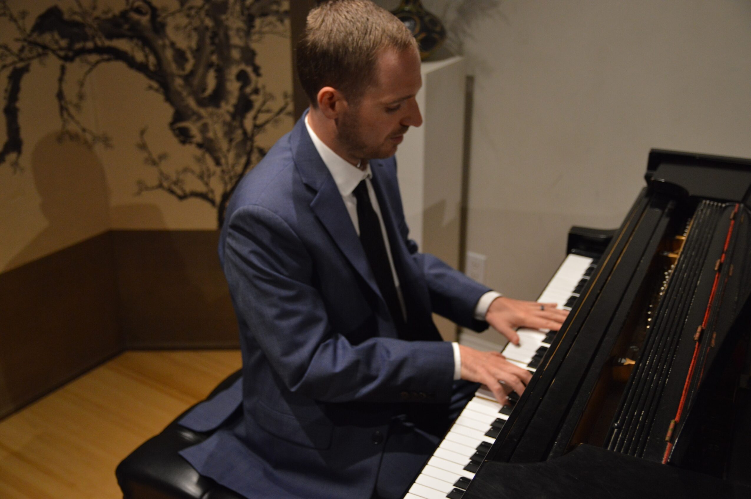 Man in a suit playing a grand piano indoors, with a tree mural on the wall behind him.