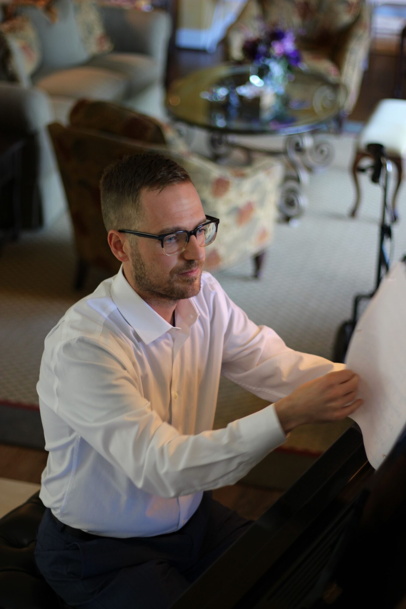 Man with glasses in a white shirt playing a piano in a cluttered room.