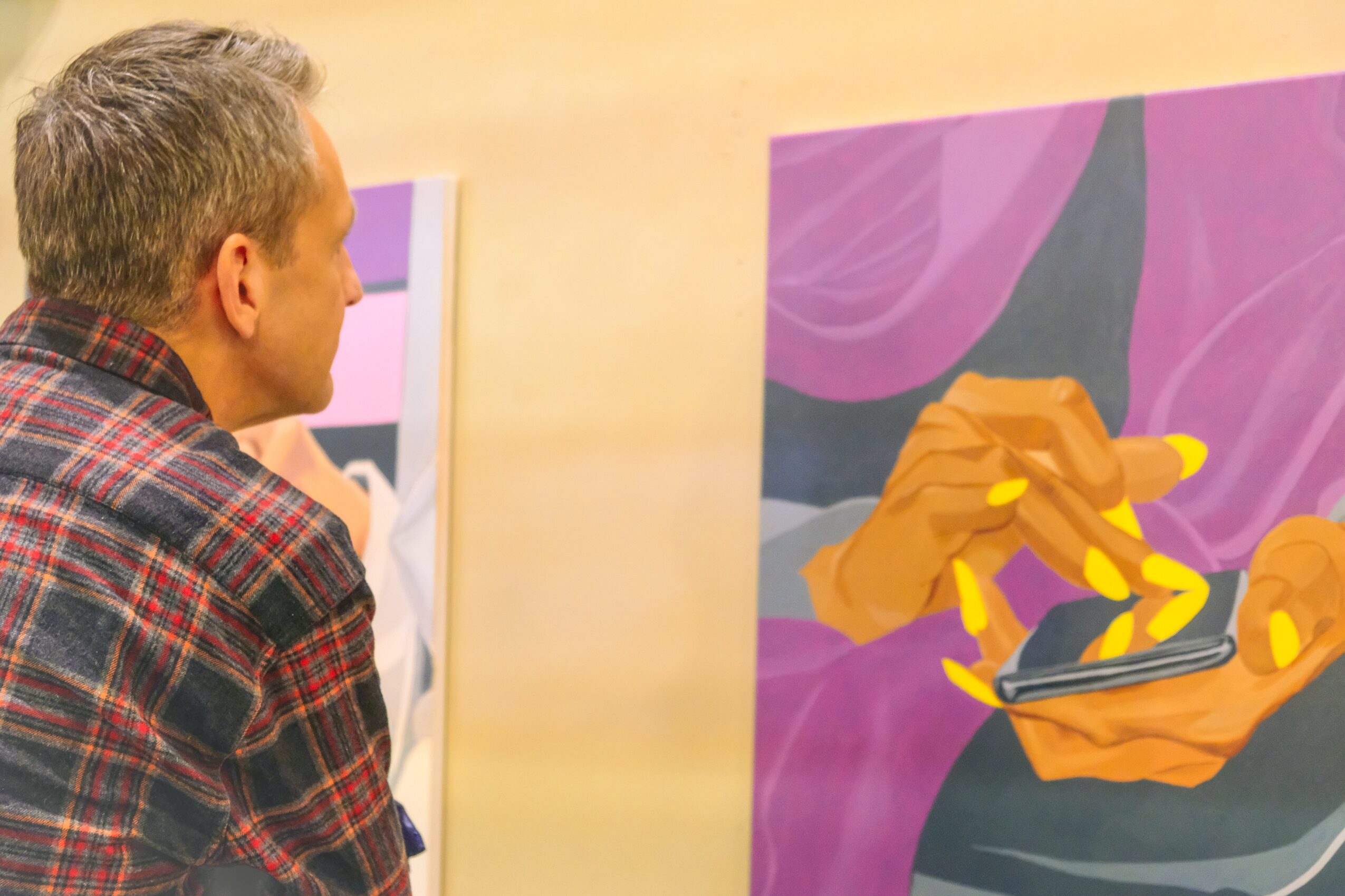 A man with gray hair wearing a plaid shirt looks at colorful abstract paintings on a wall.
