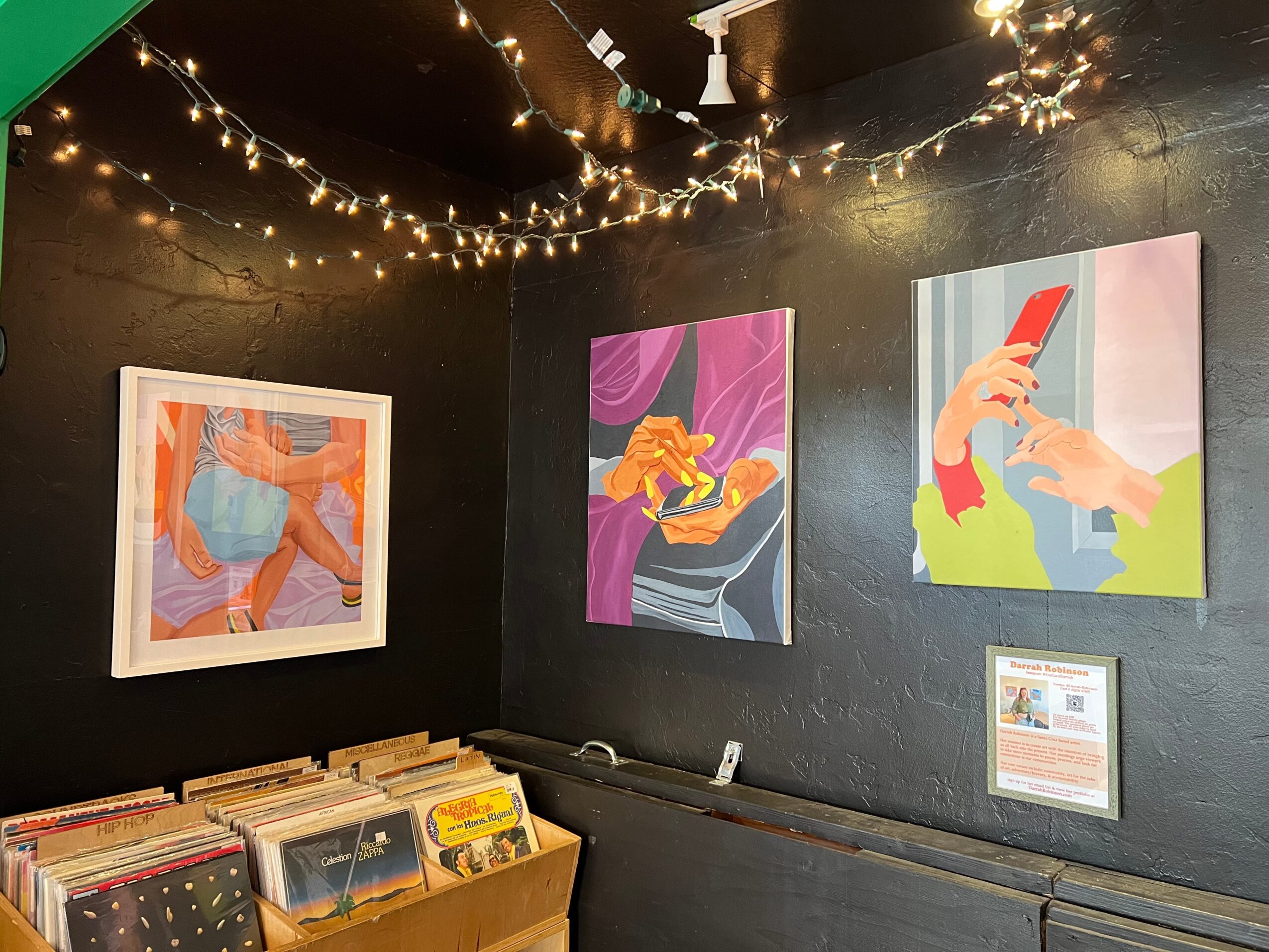 Three colorful paintings on a black wall in an art gallery, with string lights overhead and a table of vinyl records below.