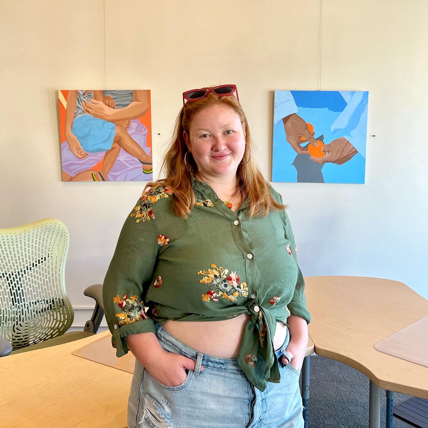 Young woman with red hair and sunglasses on her head, smiling, wearing a green floral shirt and jeans, standing in a room with artwork on the wall.