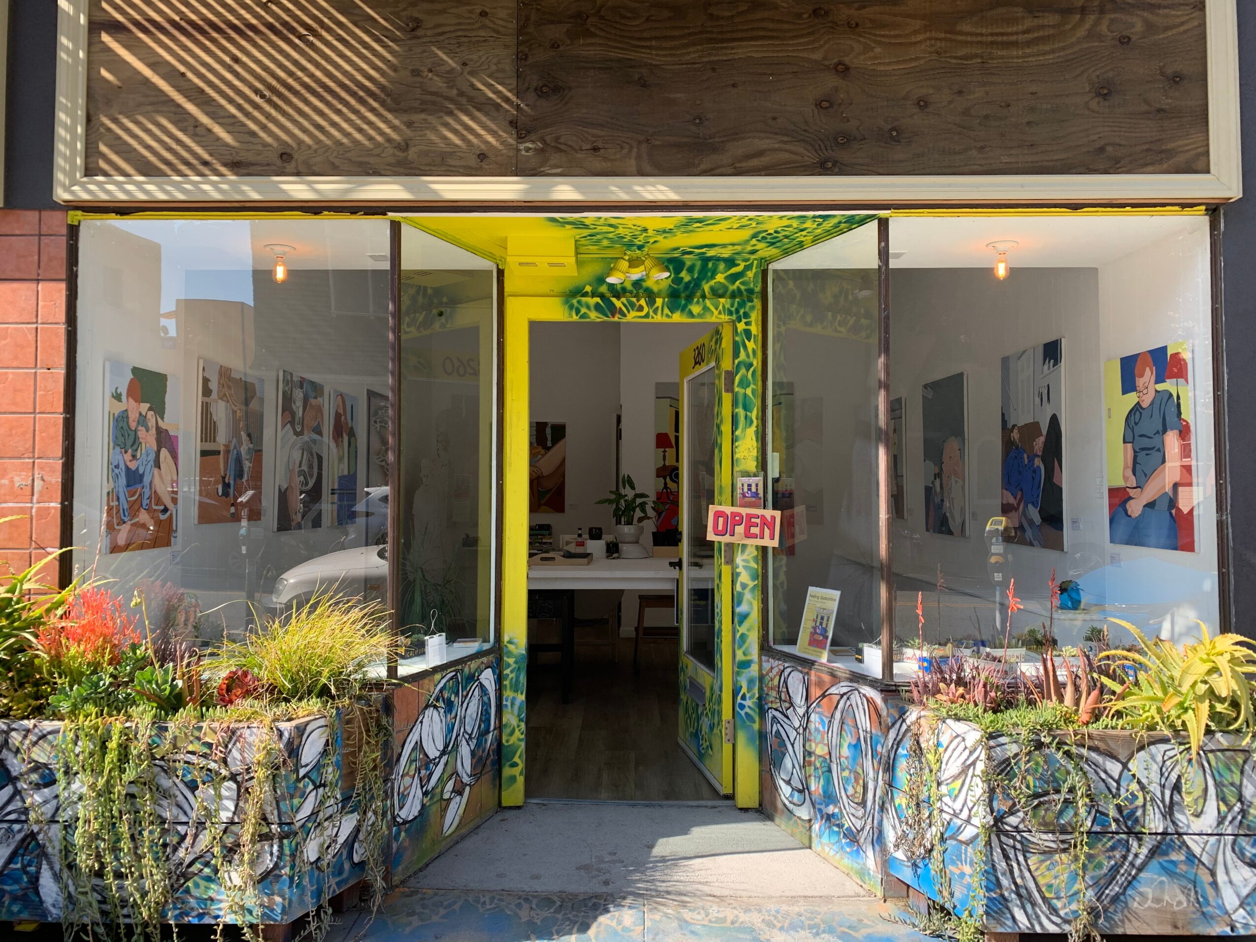 Gallery entrance with open glass doors, colorful artwork inside, plants outside, and an 'Open' sign, viewed from outside.