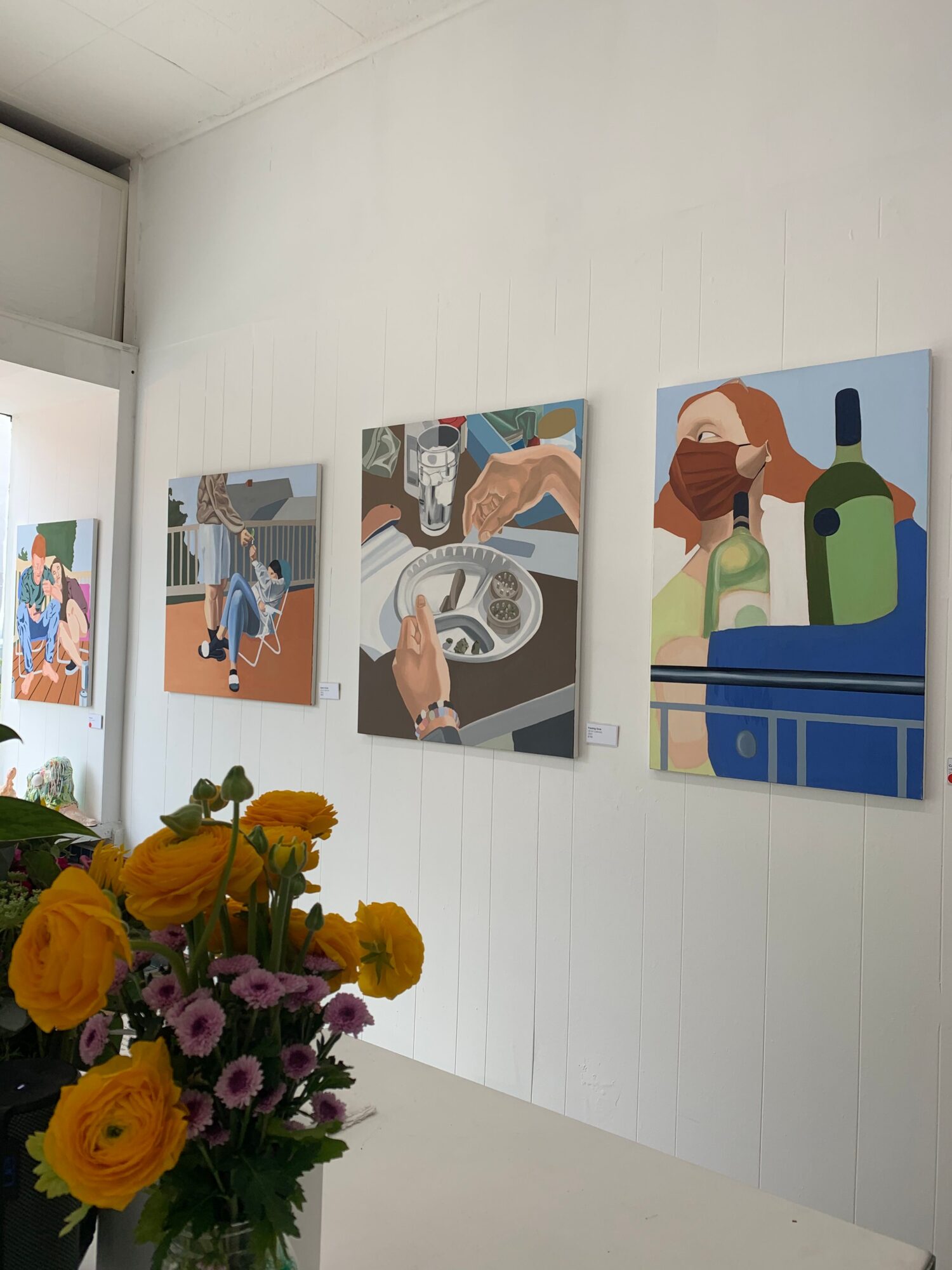 Four paintings on a white wall, with a bouquet of yellow and purple flowers in the foreground.