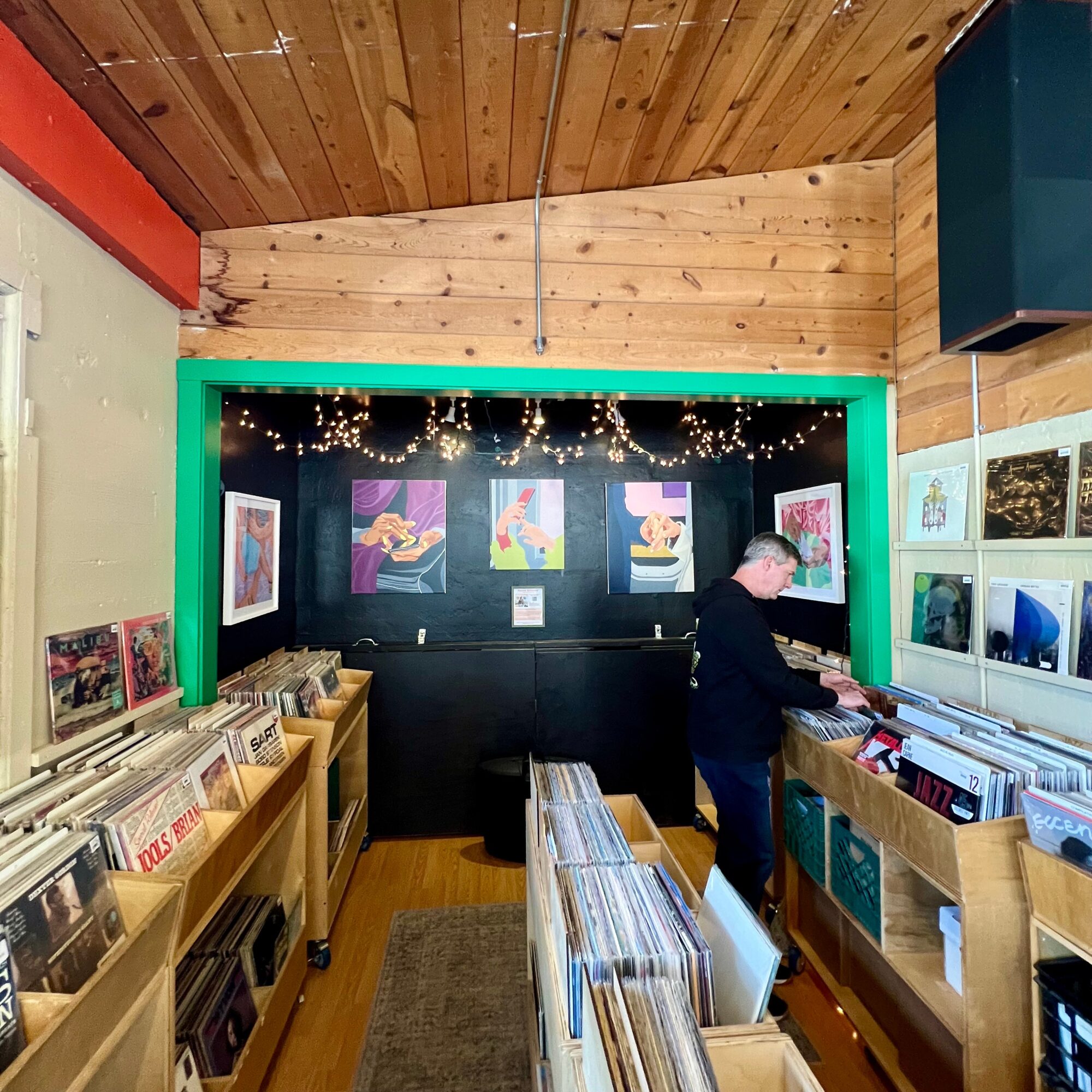 Person browsing vinyl records in a store with black wall, string lights, and colorful artwork, wooden ceiling and shelves.