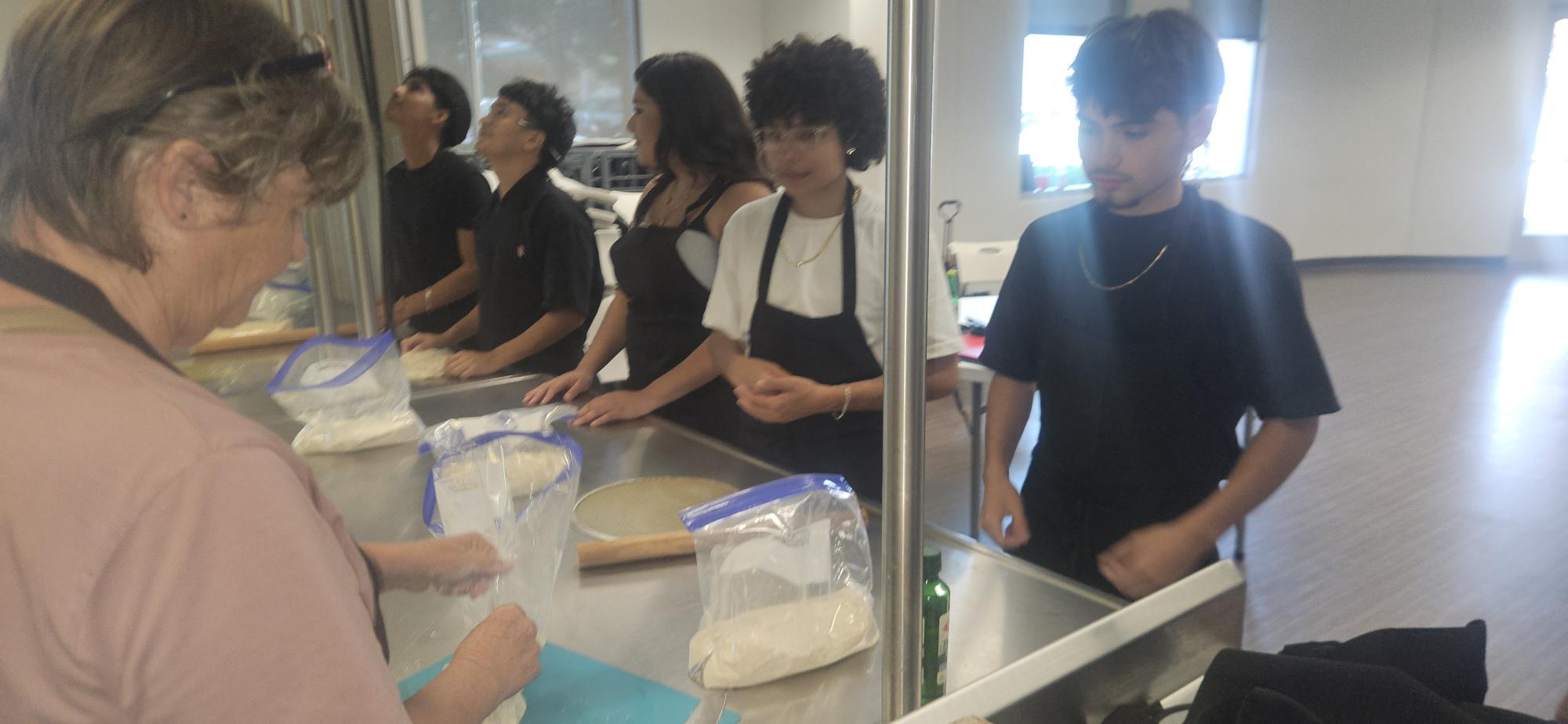 People in line at a counter, with a woman serving and others waiting, in a bright room.