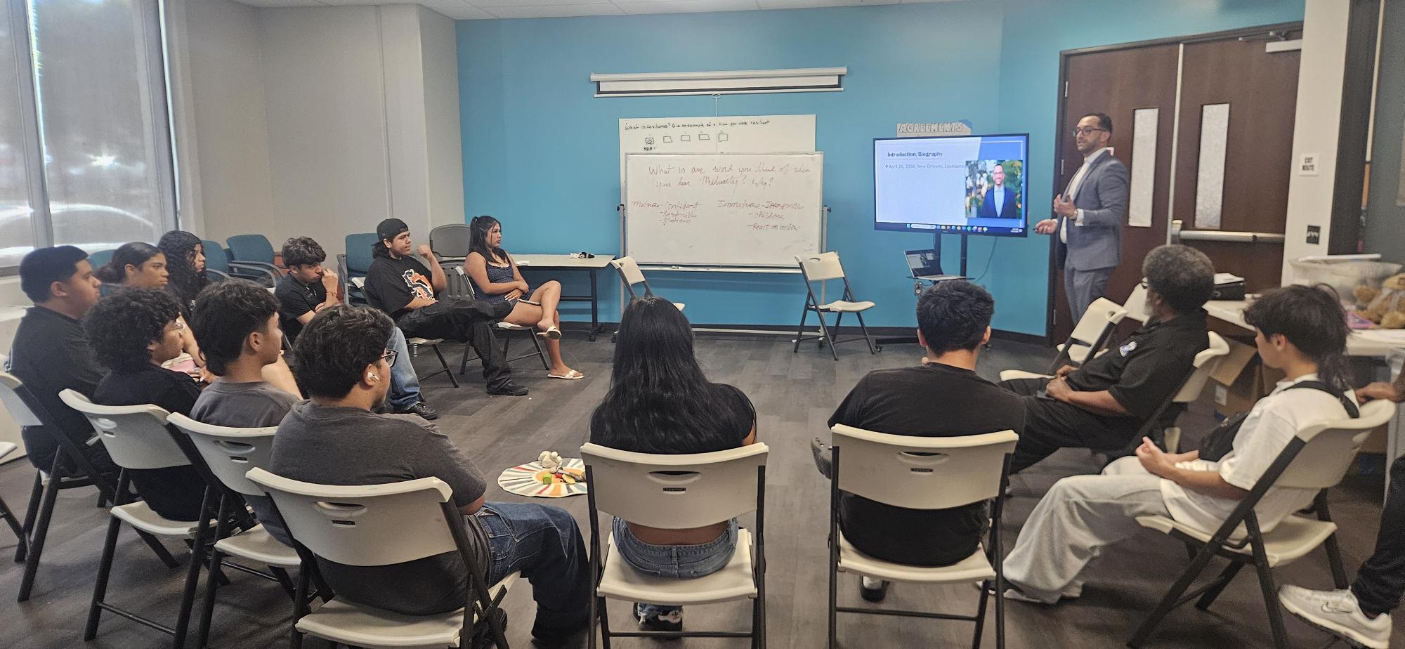 Group of people seated in a room, listening to a presenter near a whiteboard and screen.