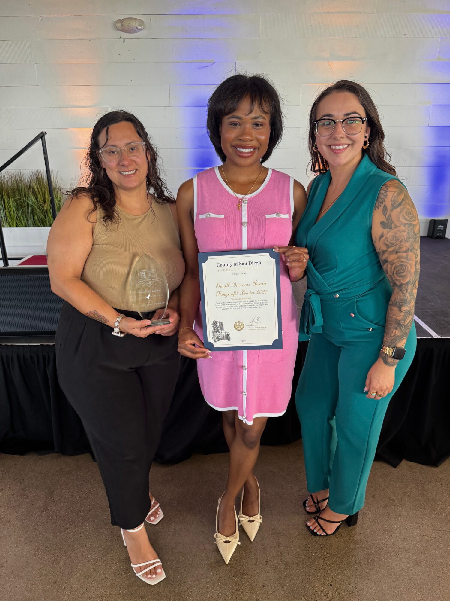 Three women standing together, one holding a framed certificate, smiling, in front of a light-colored wall with blue lighting.