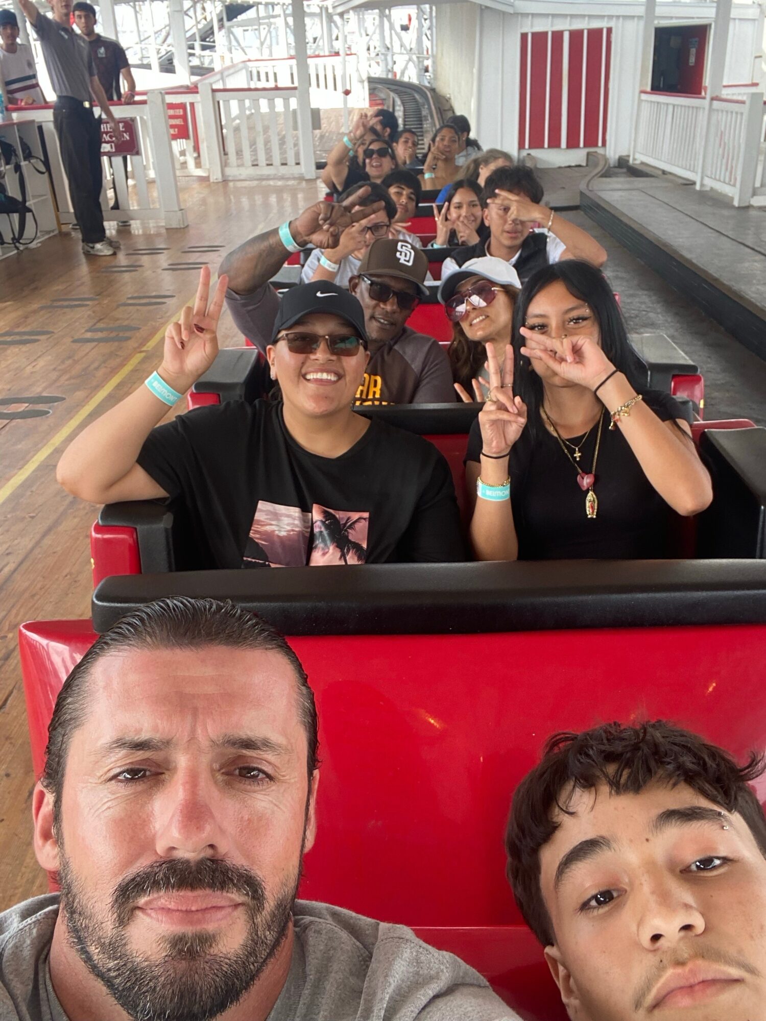 People riding a roller coaster, smiling and making peace signs, with a wooden floor and white railing in the background.