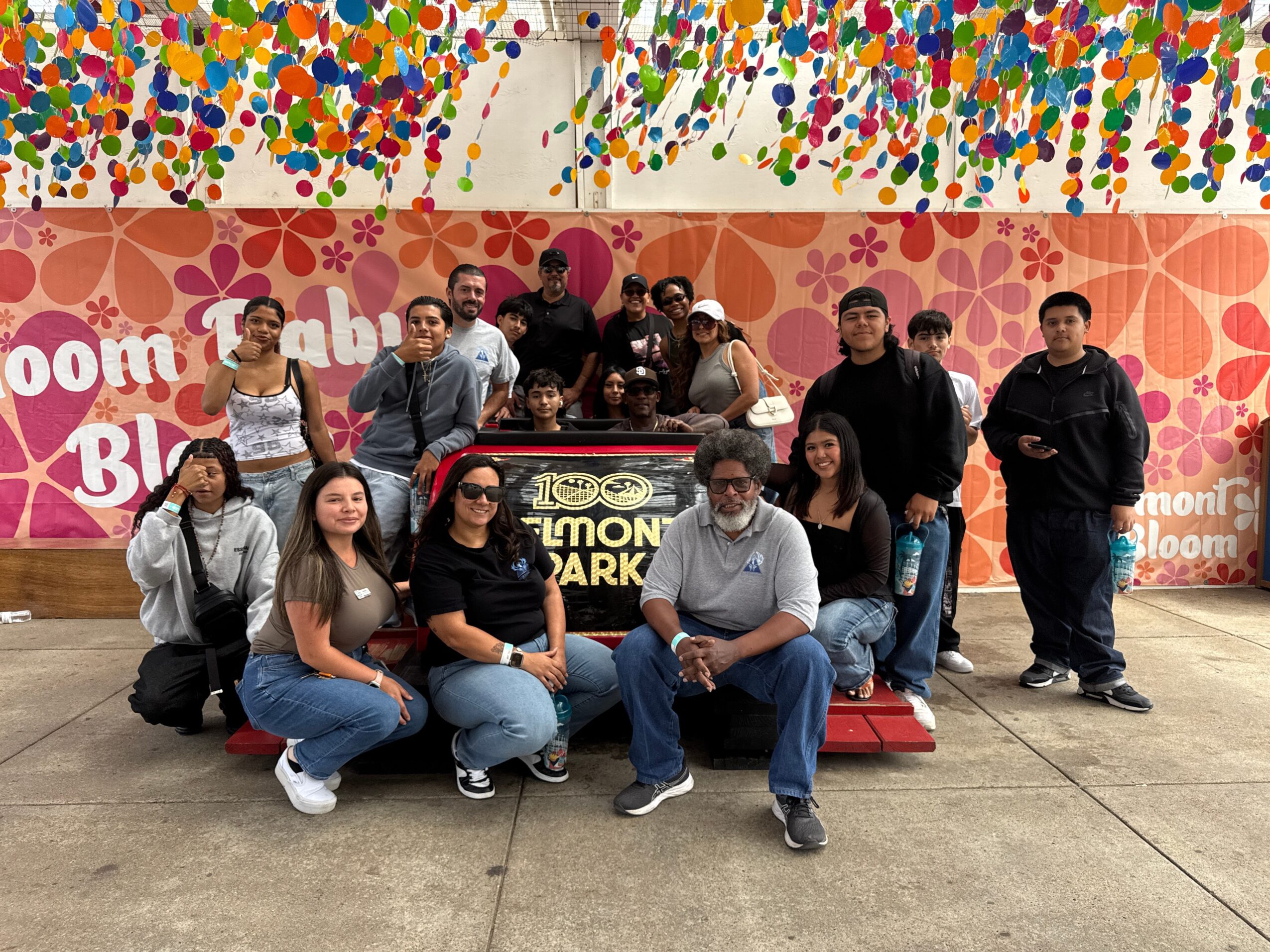 Group of people posing in front of a colorful, decorated backdrop with pink and orange patterns and multicolored hanging decorations.