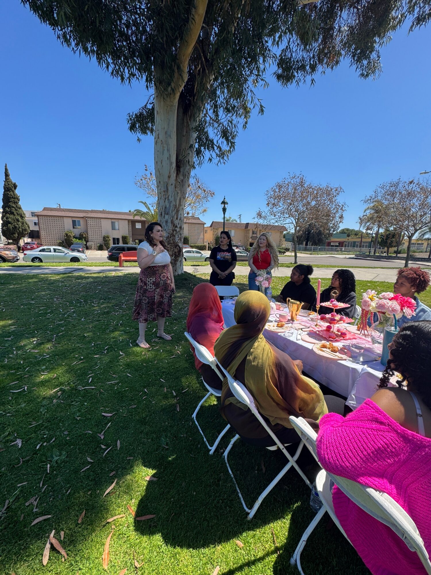 Group of women gathered outdoors under a large tree, some standing and some seated at a table with pink decorations, on a sunny day.