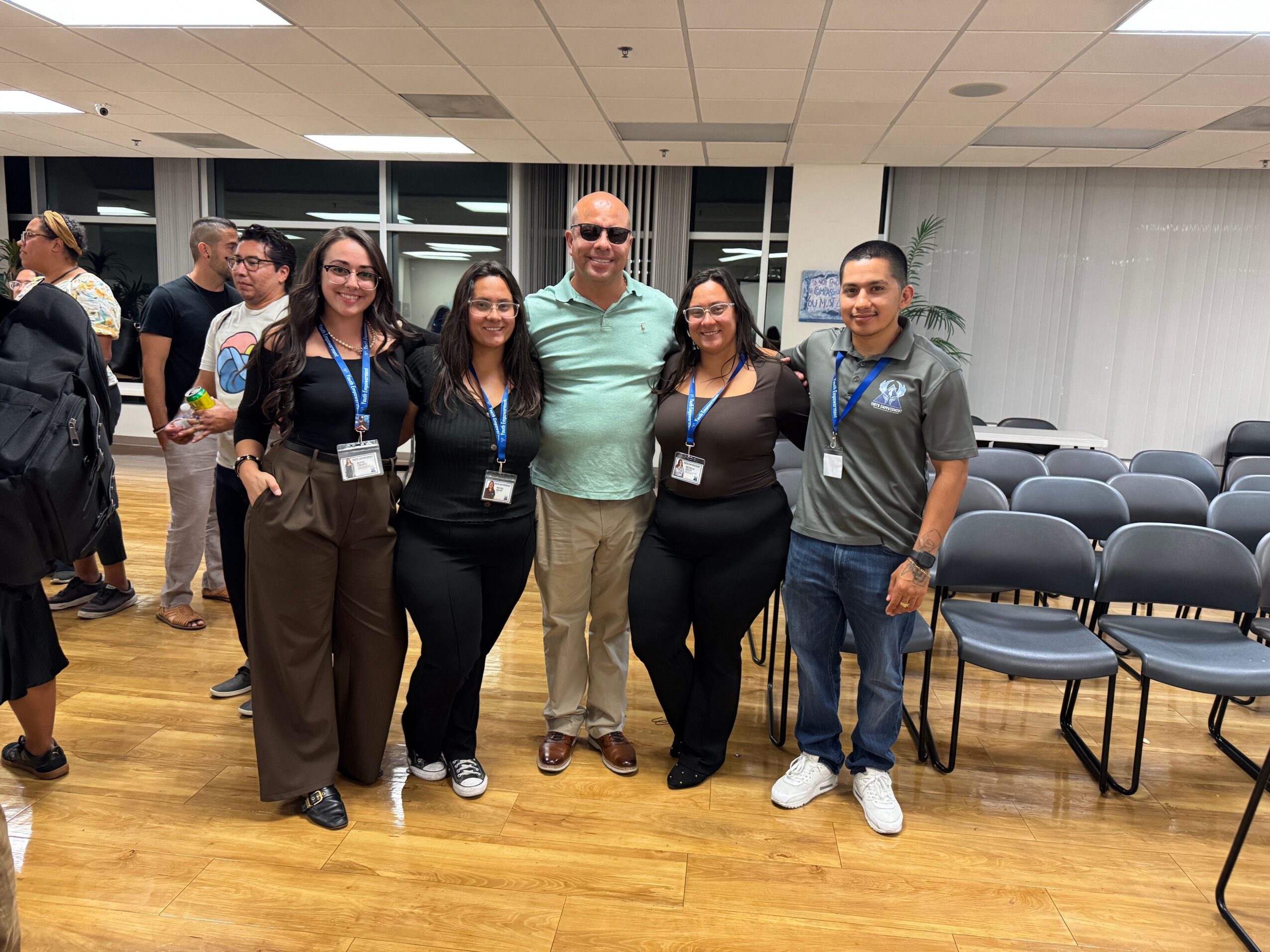 Group of five people standing indoors, smiling, with chairs and other people in the background.