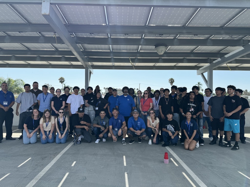 Group of people standing and sitting under a metal roof structure, with a parking lot and trees in the background.