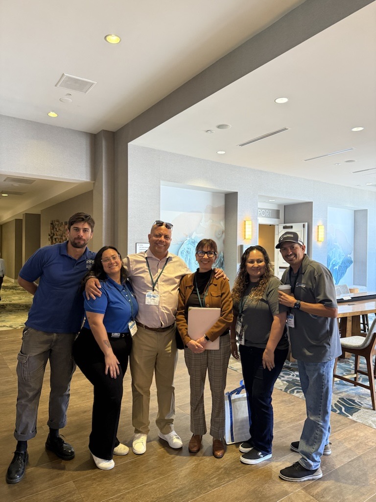 Six people standing together indoors, smiling, with a modern interior background, some holding folders or bags.