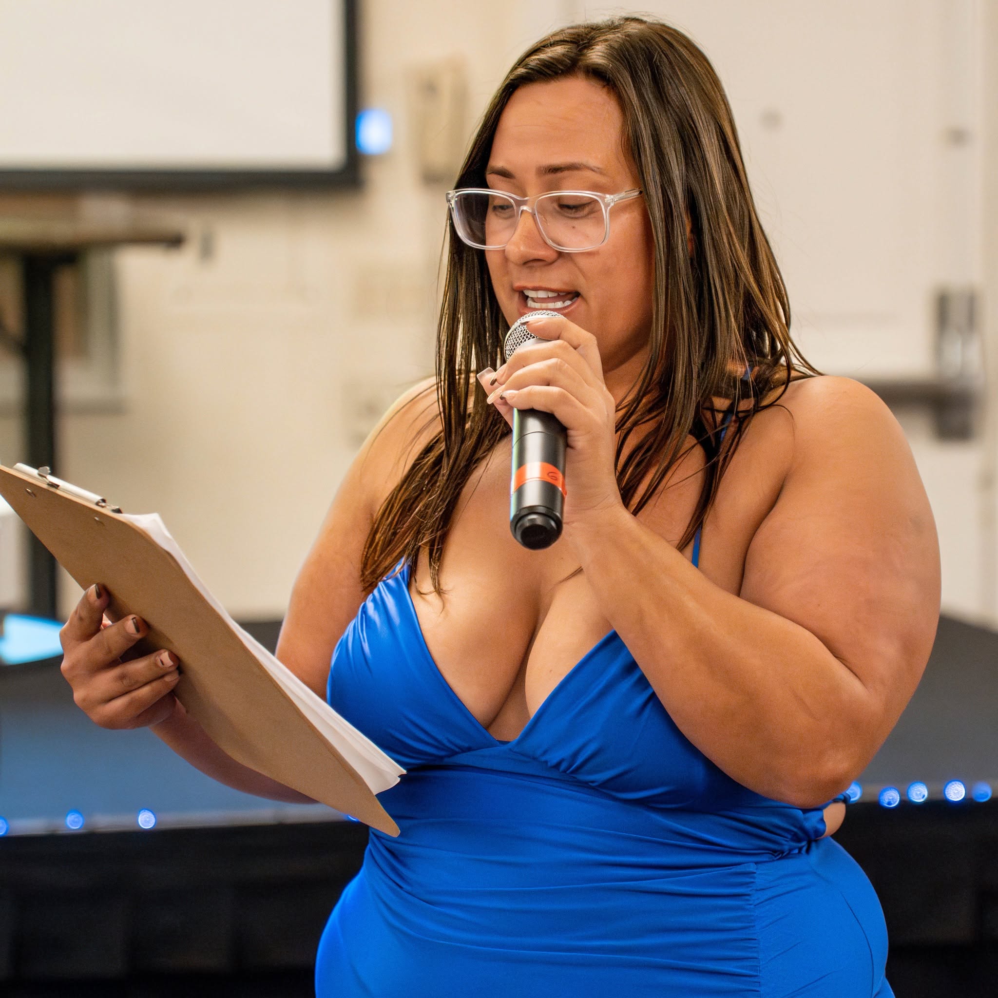 Woman in blue dress speaking into microphone and reading from paper, glasses, indoor setting
