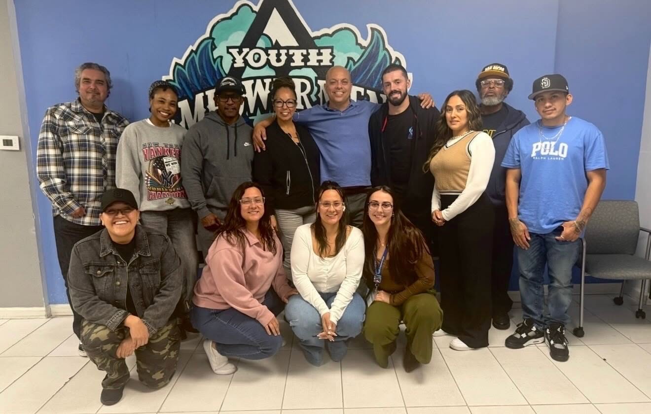 Group of 13 people posing indoors in front of a Youth Wilderness sign, some standing and some kneeling, smiling.
