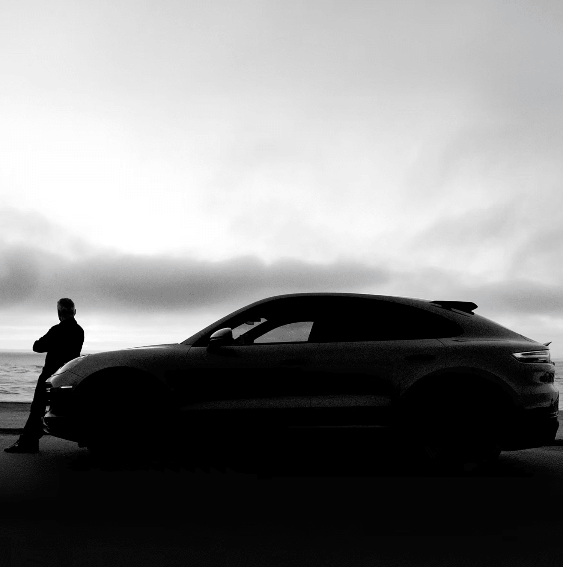 Silhouette of a person standing next to a car against a cloudy sky, in black and white.
