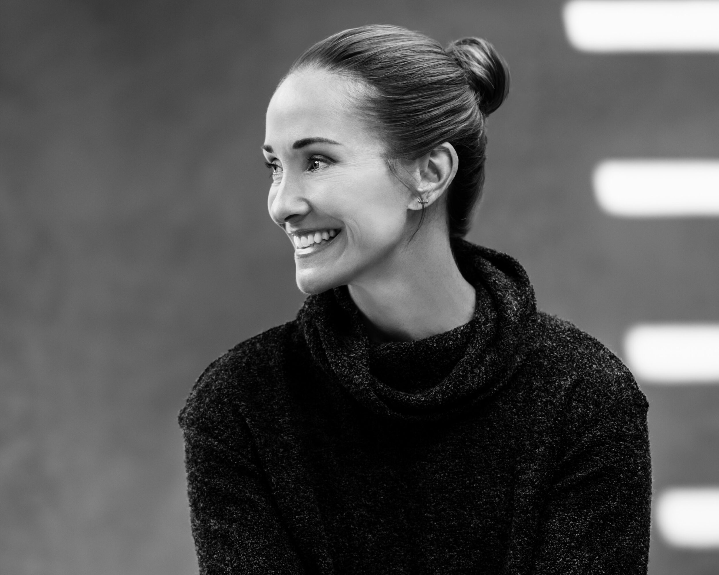 Woman smiling, hair in a bun, wearing a dark sweater, looking to the side, background with horizontal lines.