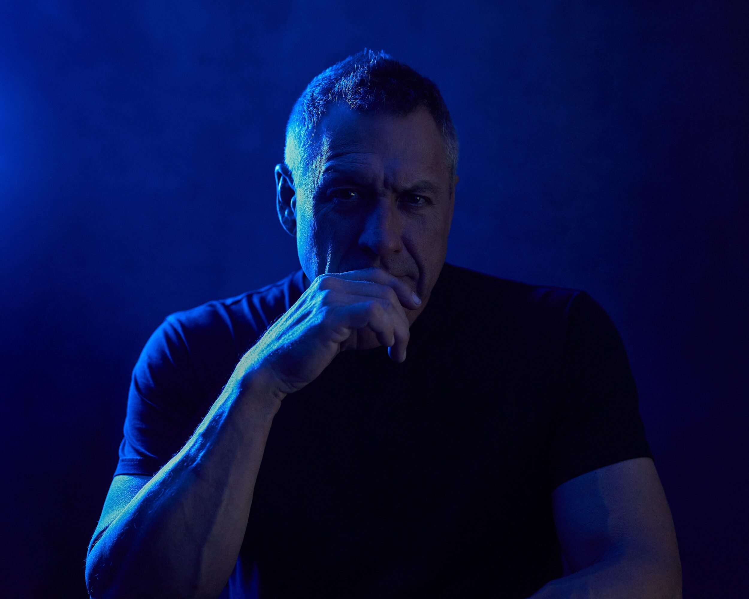 Man with short hair and a serious expression, hand near mouth, in dark blue lighting.